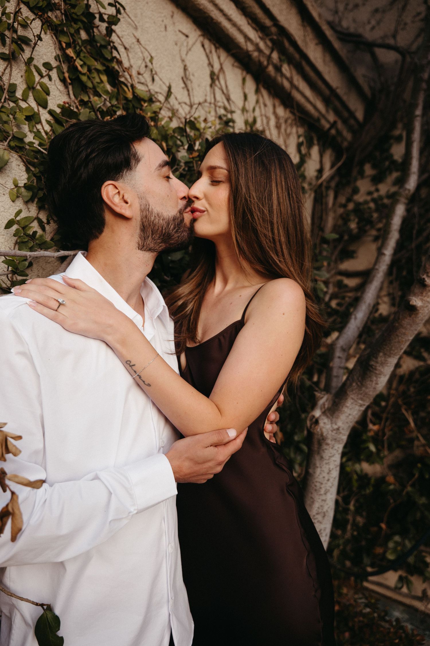 A couple shares a romantic kiss while embracing outdoors, woman in dark slip dress and man in white shirt.