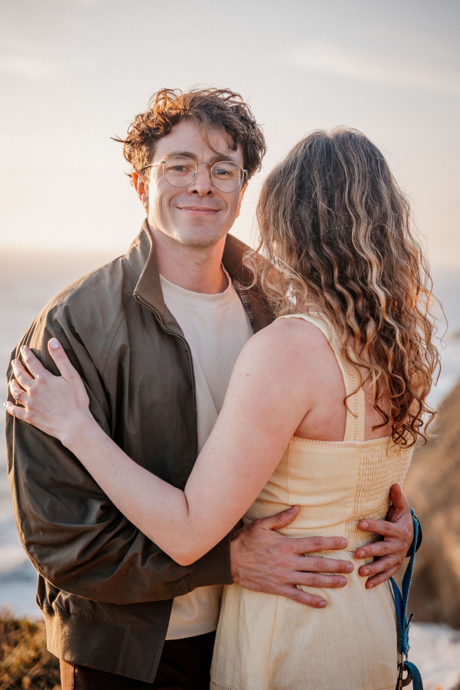 Happy man in glasses and olive jacket embracing woman at sunset by the ocean.