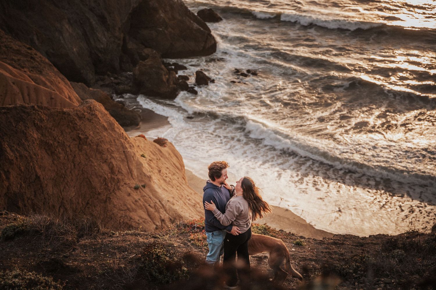 Couple embracing romantically on rocky coastal cliffs at golden sunset with ocean waves crashing below.