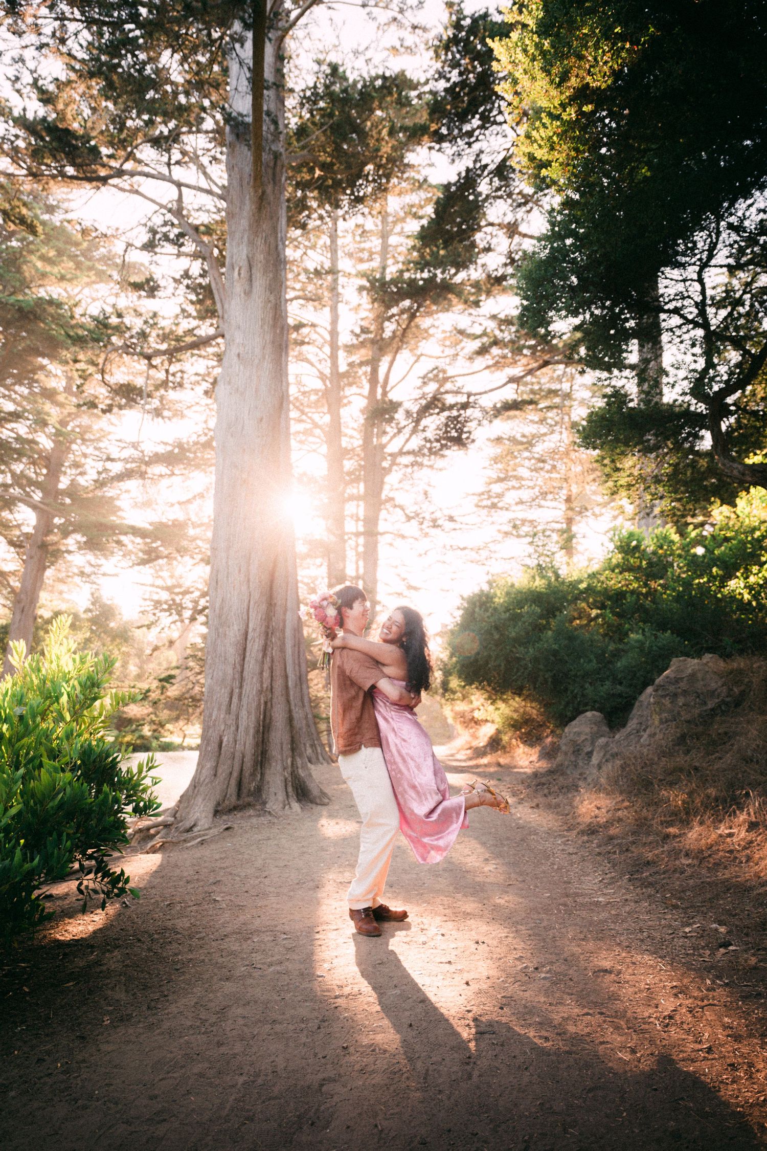 Couple embracing and spinning in golden sunlight among tall trees on a forest path.