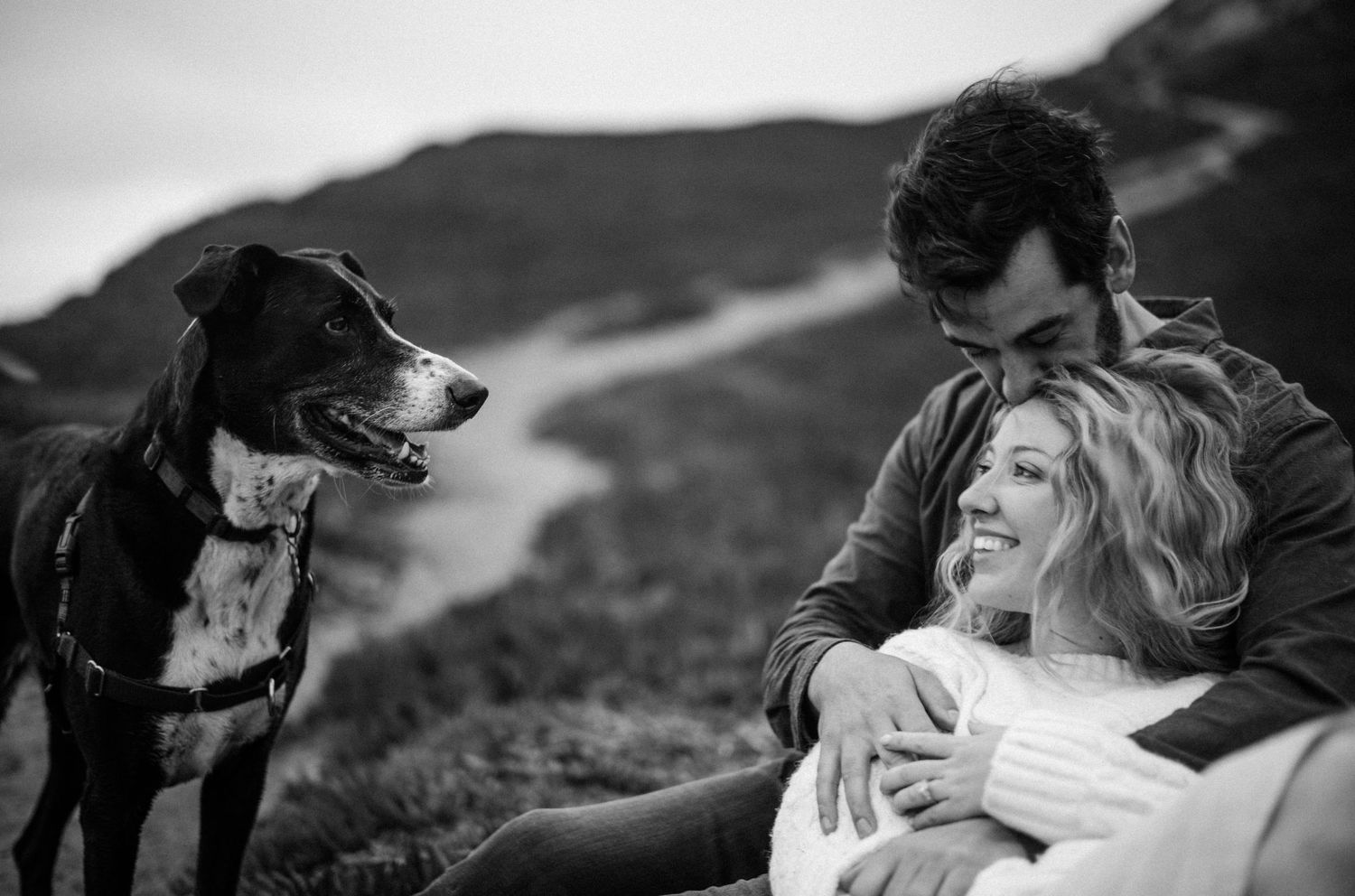 Black and white photo of couple sitting with happy dog on scenic hillside overlook.