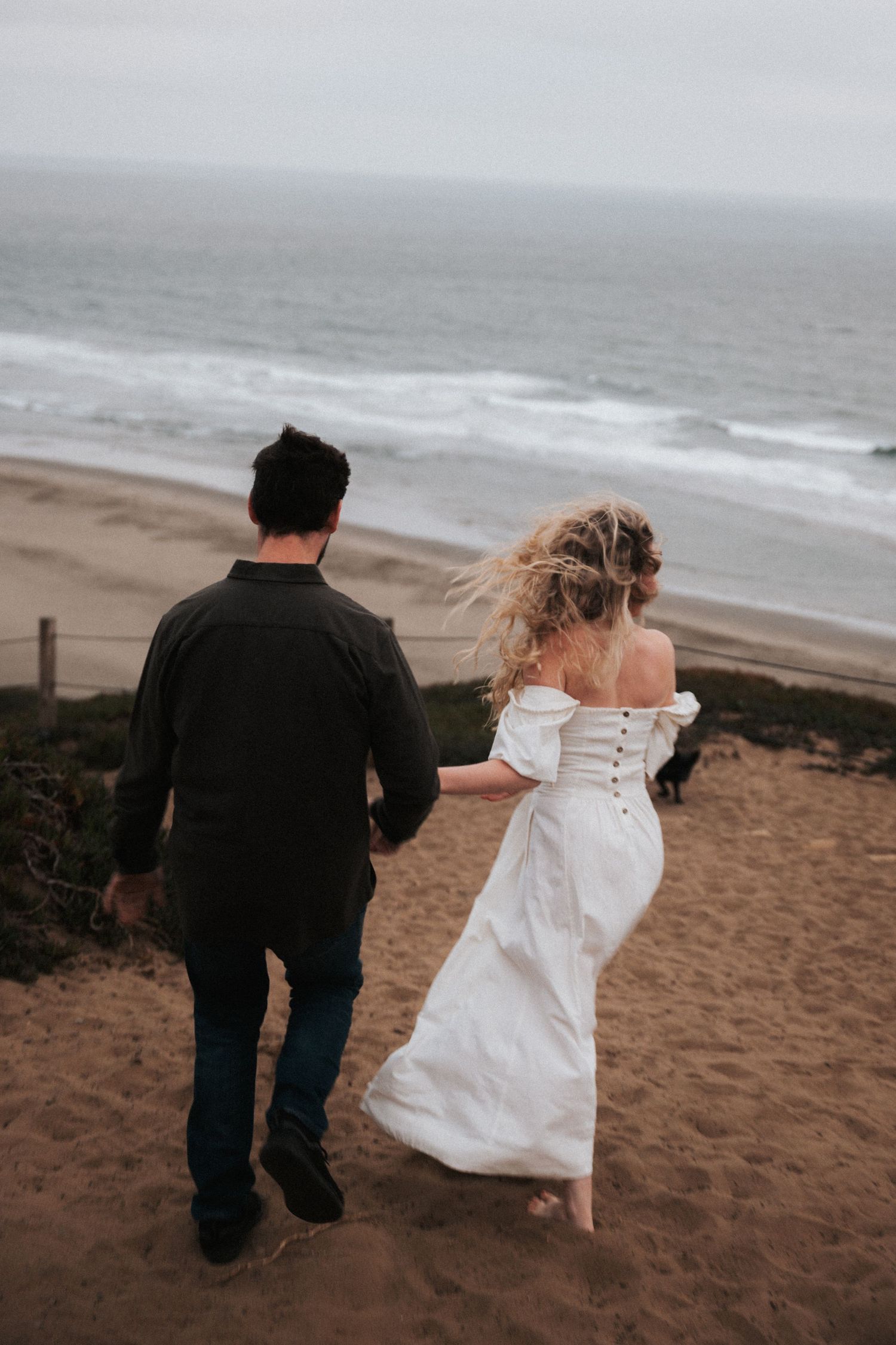 Couple holding hands walking toward beach on sandy cliff, woman in white off-shoulder dress, man in dark shirt.
