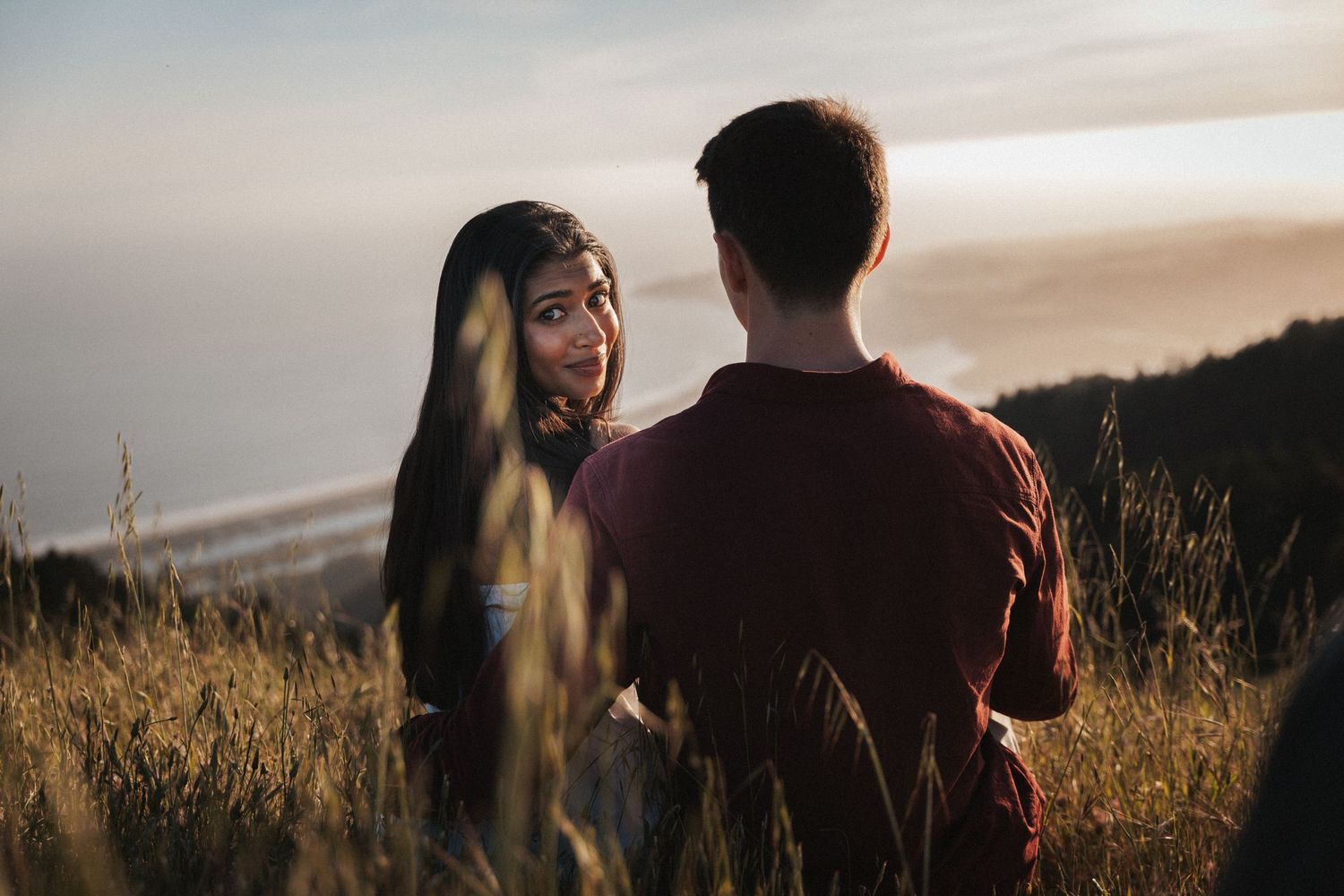 A young couple shares a romantic moment in a golden field at sunset with a scenic coastal view in the background.Photography by The Y.K. Stu