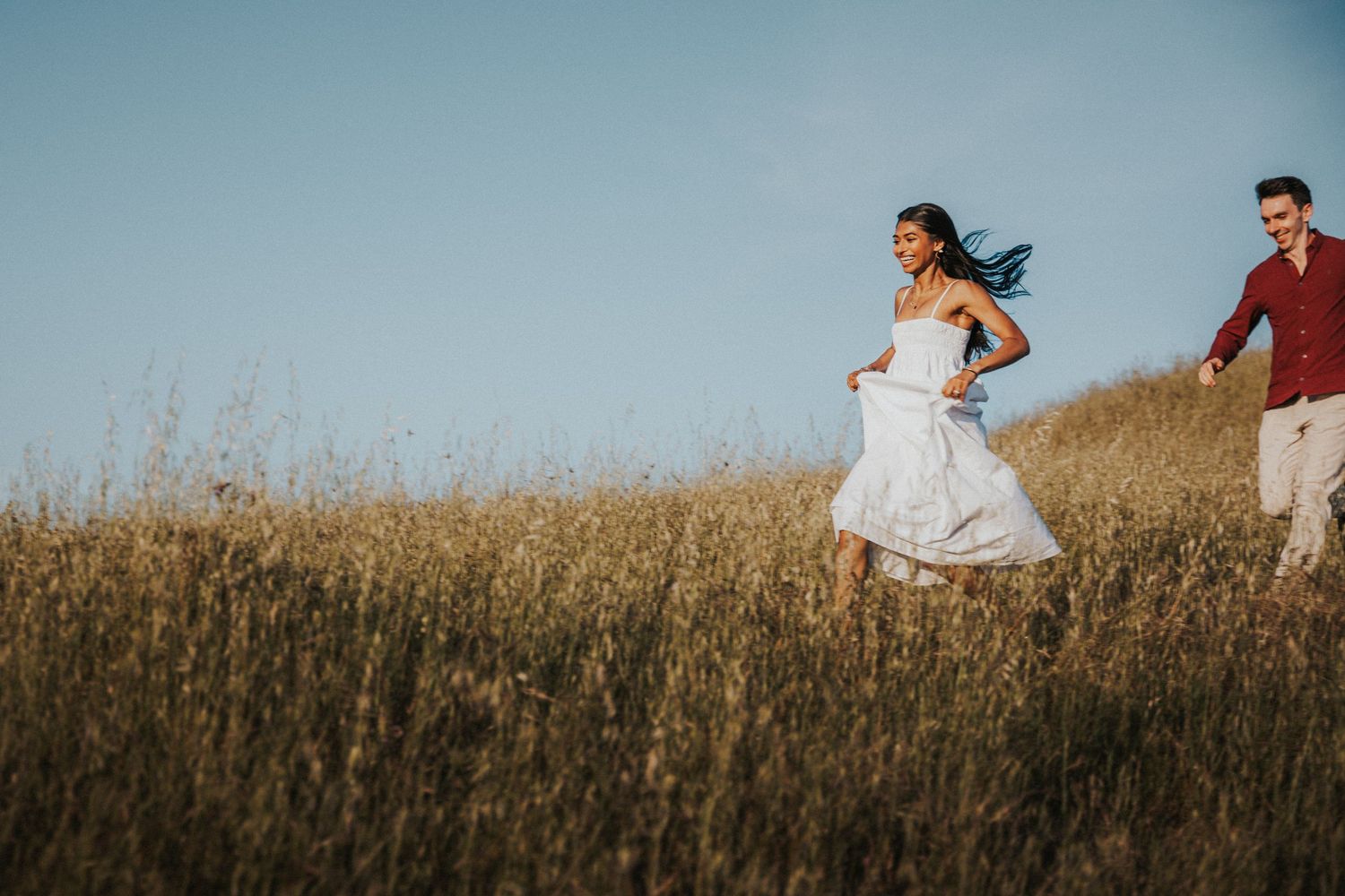 Couple running joyfully through golden wheat field under blue sky, woman in white dress, romantic outdoor photo.Photography by The Y.K. Stud
