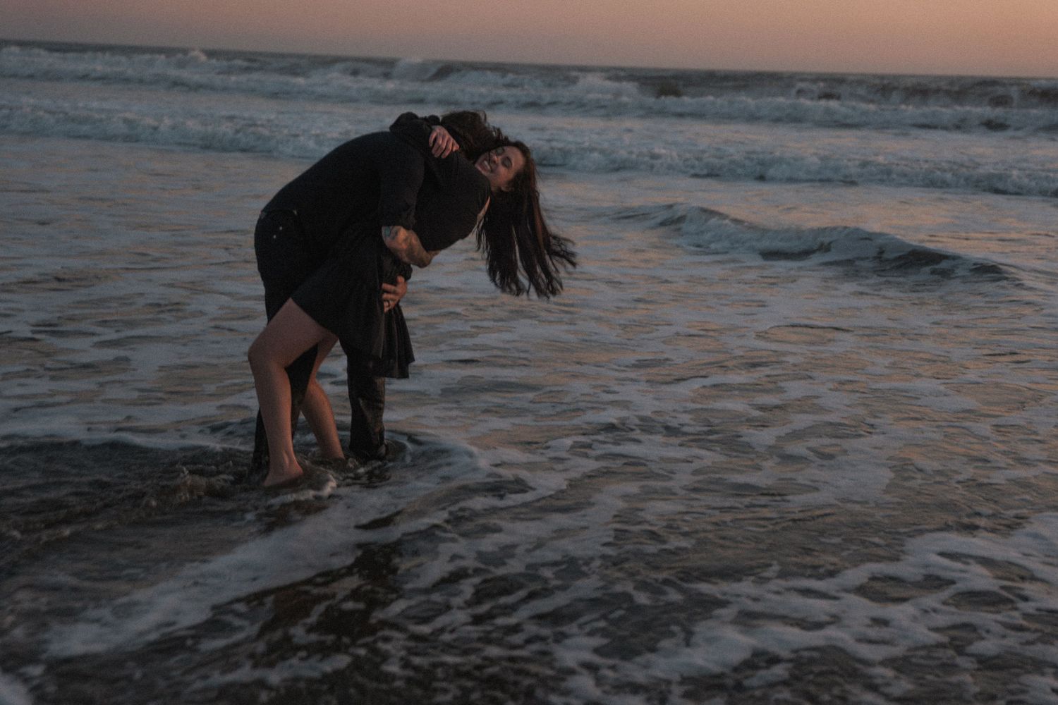 Couple embracing playfully in ocean waves at sunset on a sandy beach.