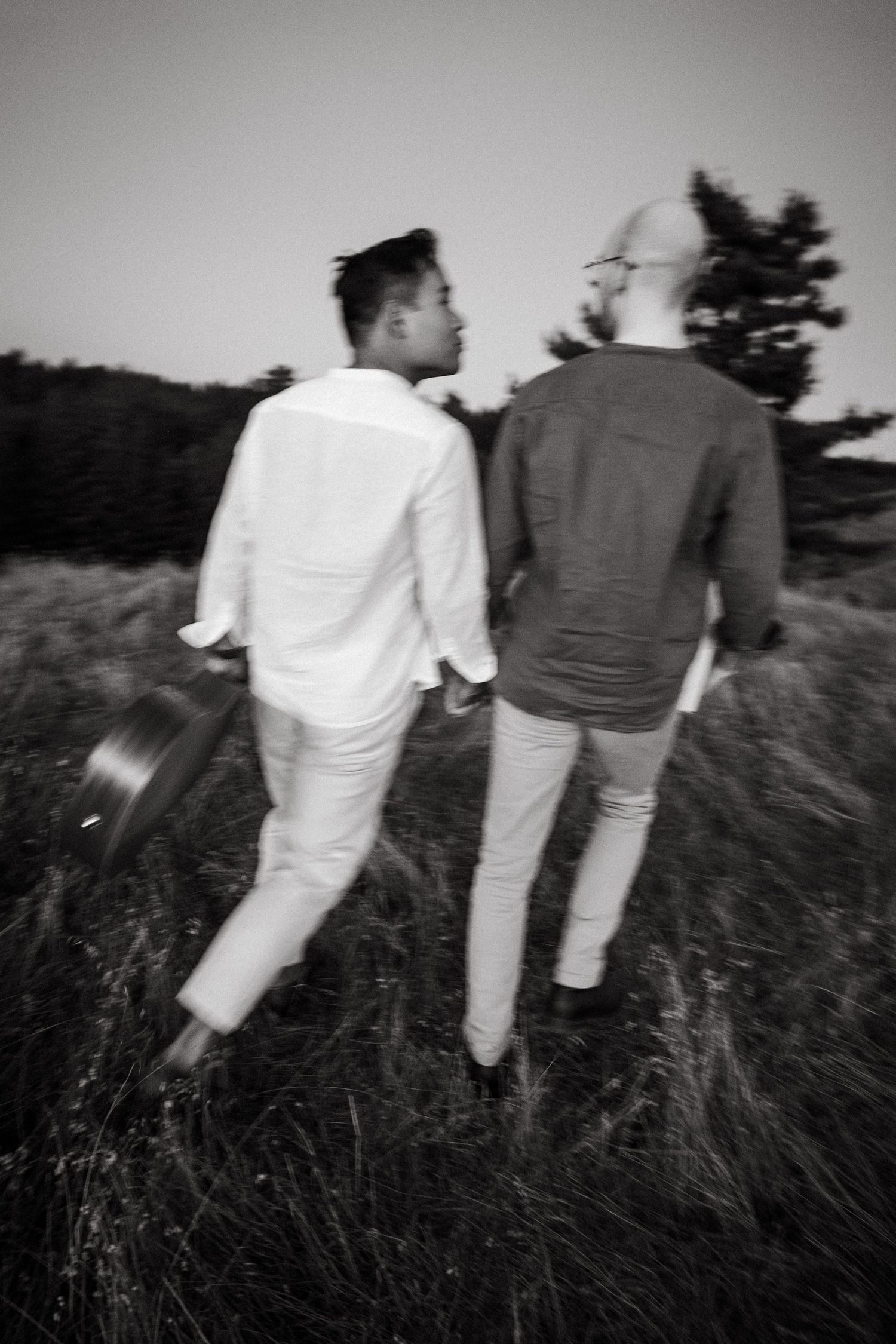 Black and white photo of two men walking through tall grass field, one carrying a guitar at dusk.