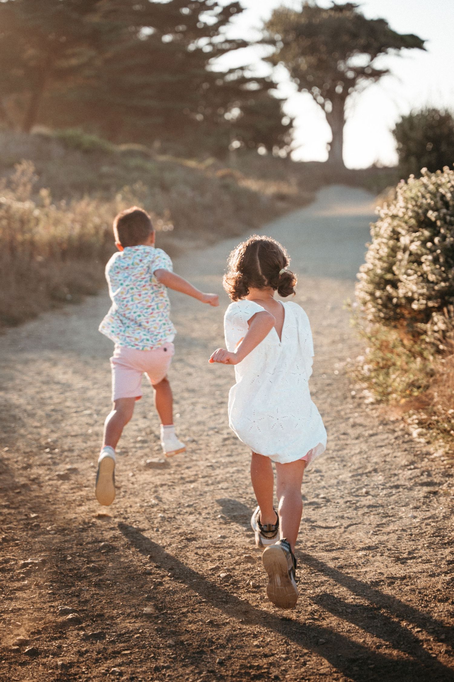 Two young children running down a sunny dirt path surrounded by trees and shrubs in golden hour light.