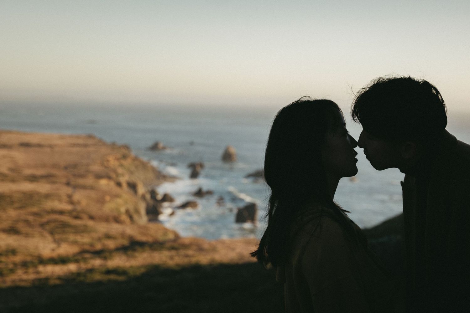 Silhouetted couple leaning in for a kiss on a coastal cliff at sunset with ocean waves and rocky shoreline in the background.