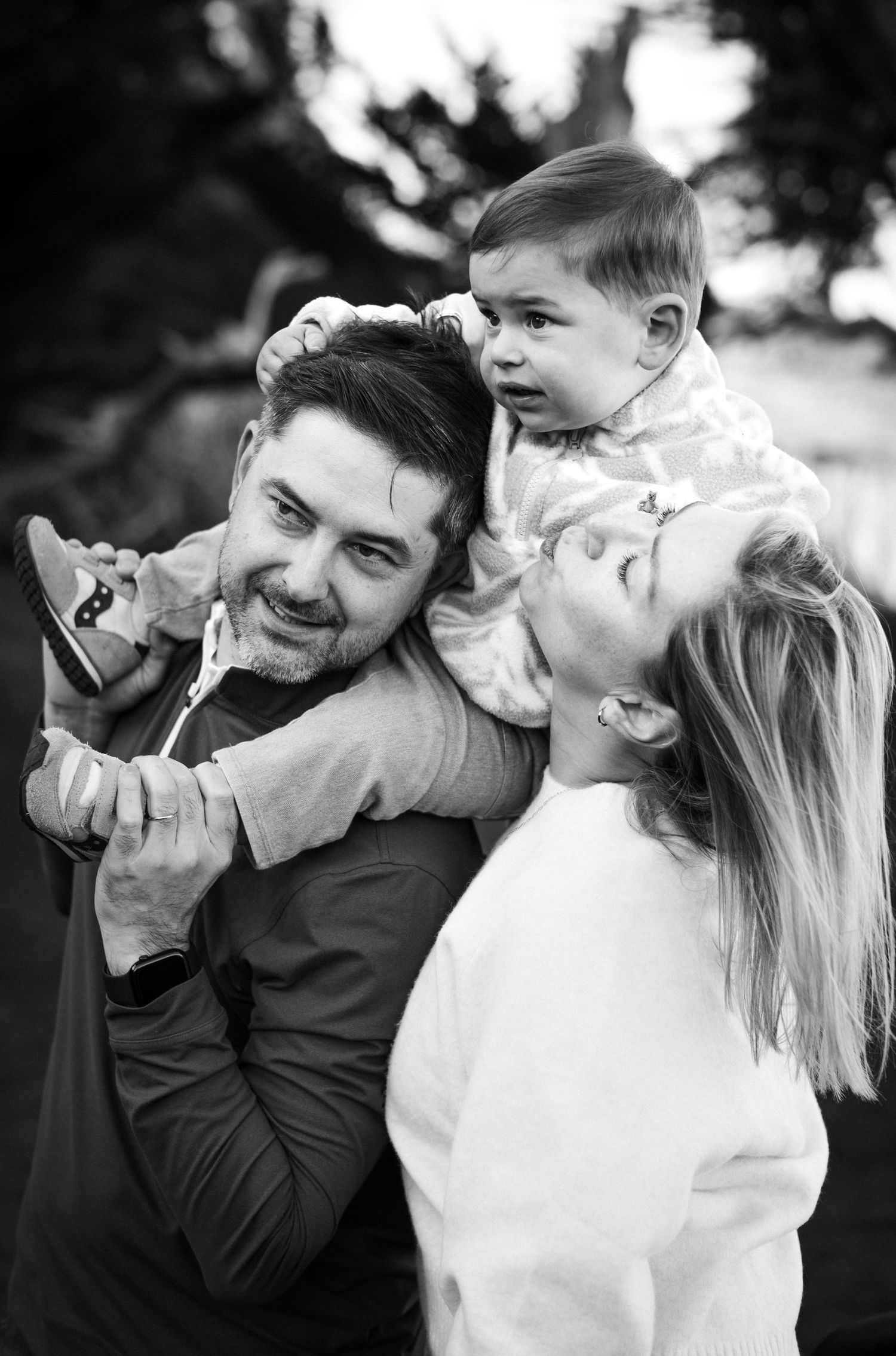 Black and white photo of a smiling couple playfully holding their happy baby outdoors in a candid family portrait.