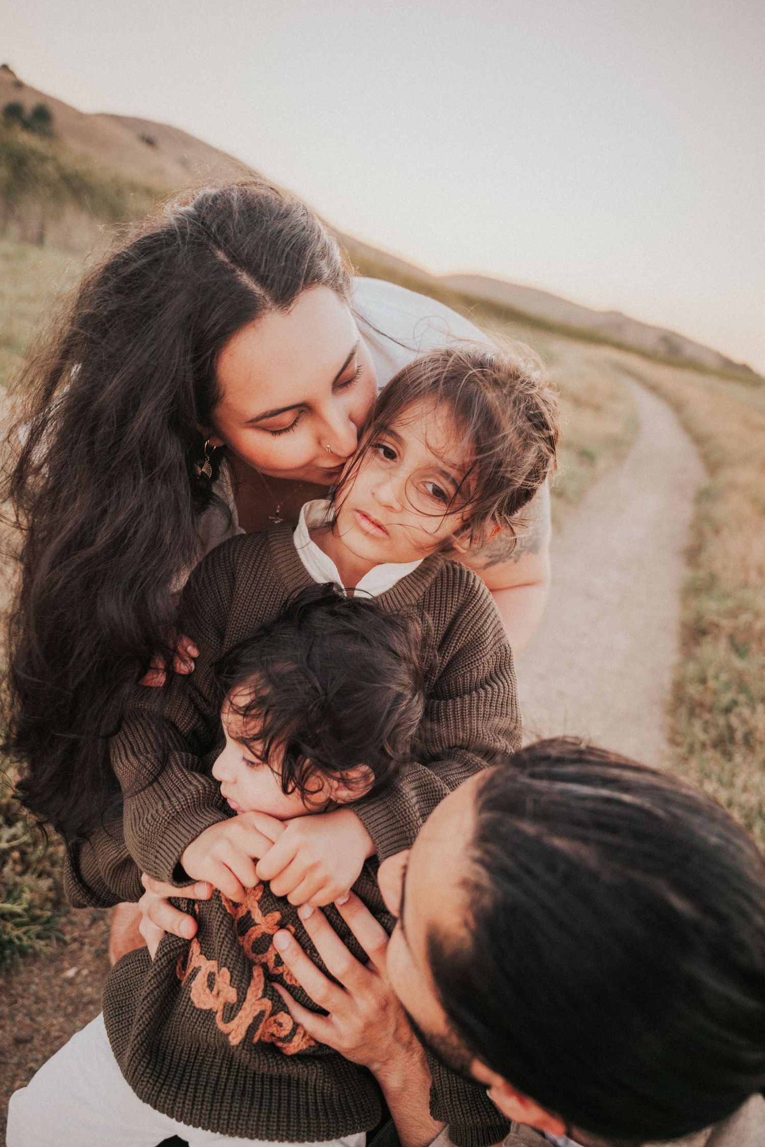 A mother tenderly kisses her young daughter while embracing both children on a scenic hillside path at sunset.