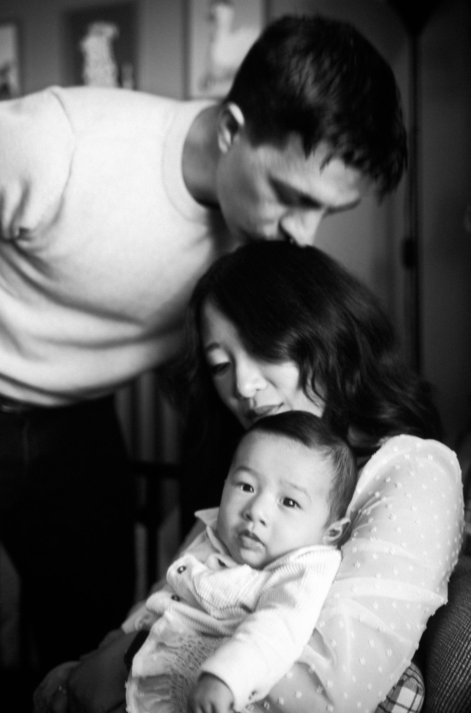 Black and white photo of a loving couple kissing their newborn baby in a tender family portrait.