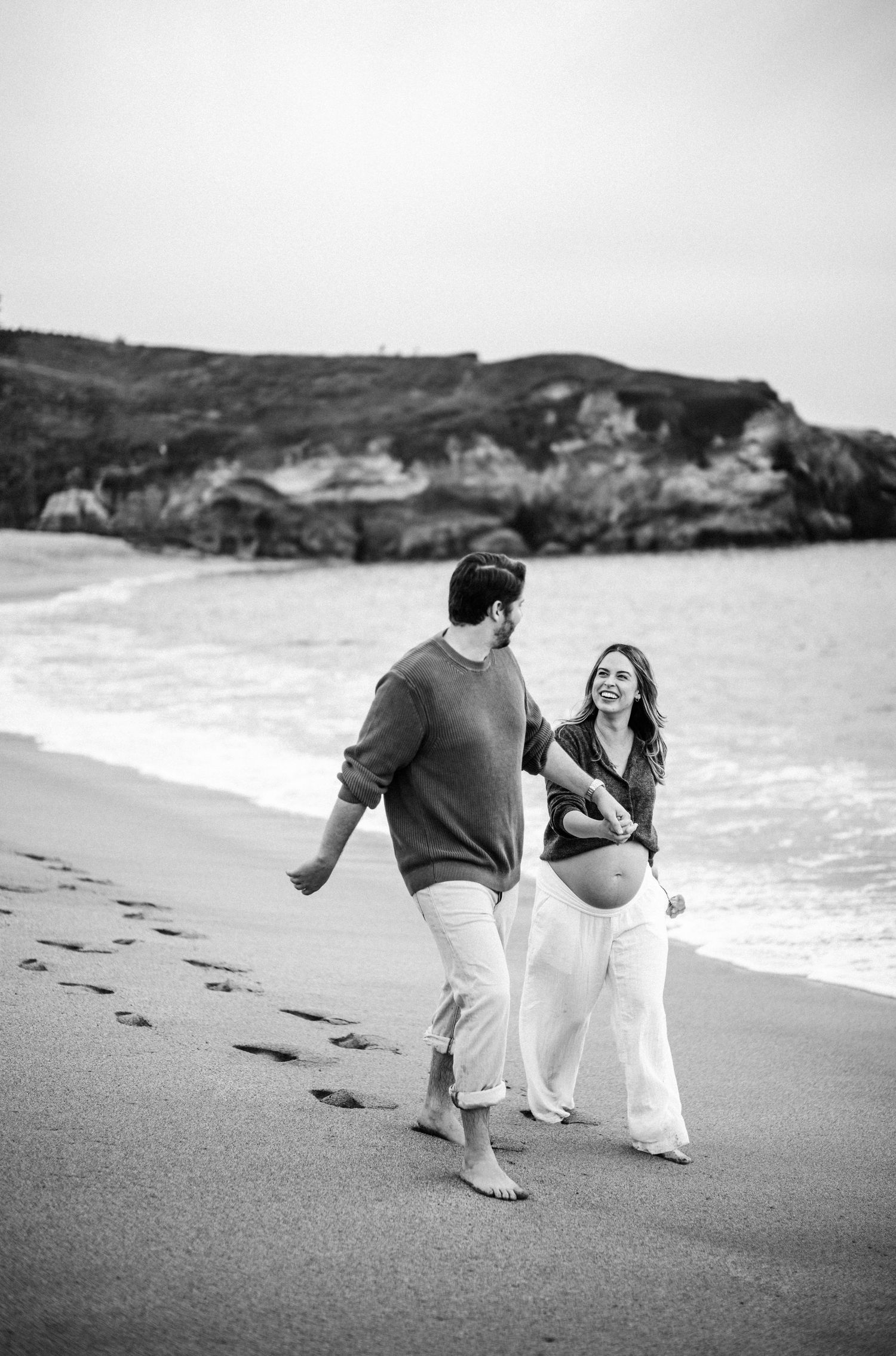 Black and white photo of a pregnant couple walking barefoot on a beach shoreline together.