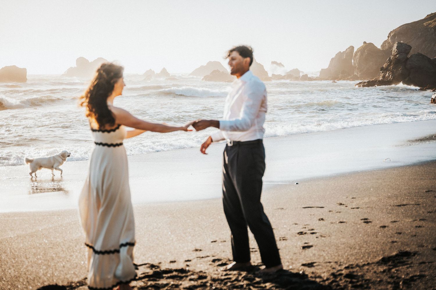 Couple holding hands on a black sand beach at sunset with a white dog and rocky ocean waves in the background.Photography by The Y.K. Studio