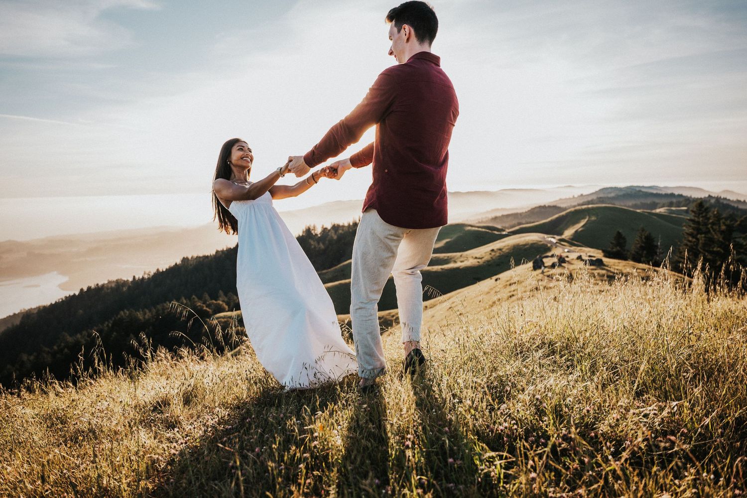 Couple dancing and holding hands on scenic hilltop meadow at golden hour with misty mountain valley backdrop.