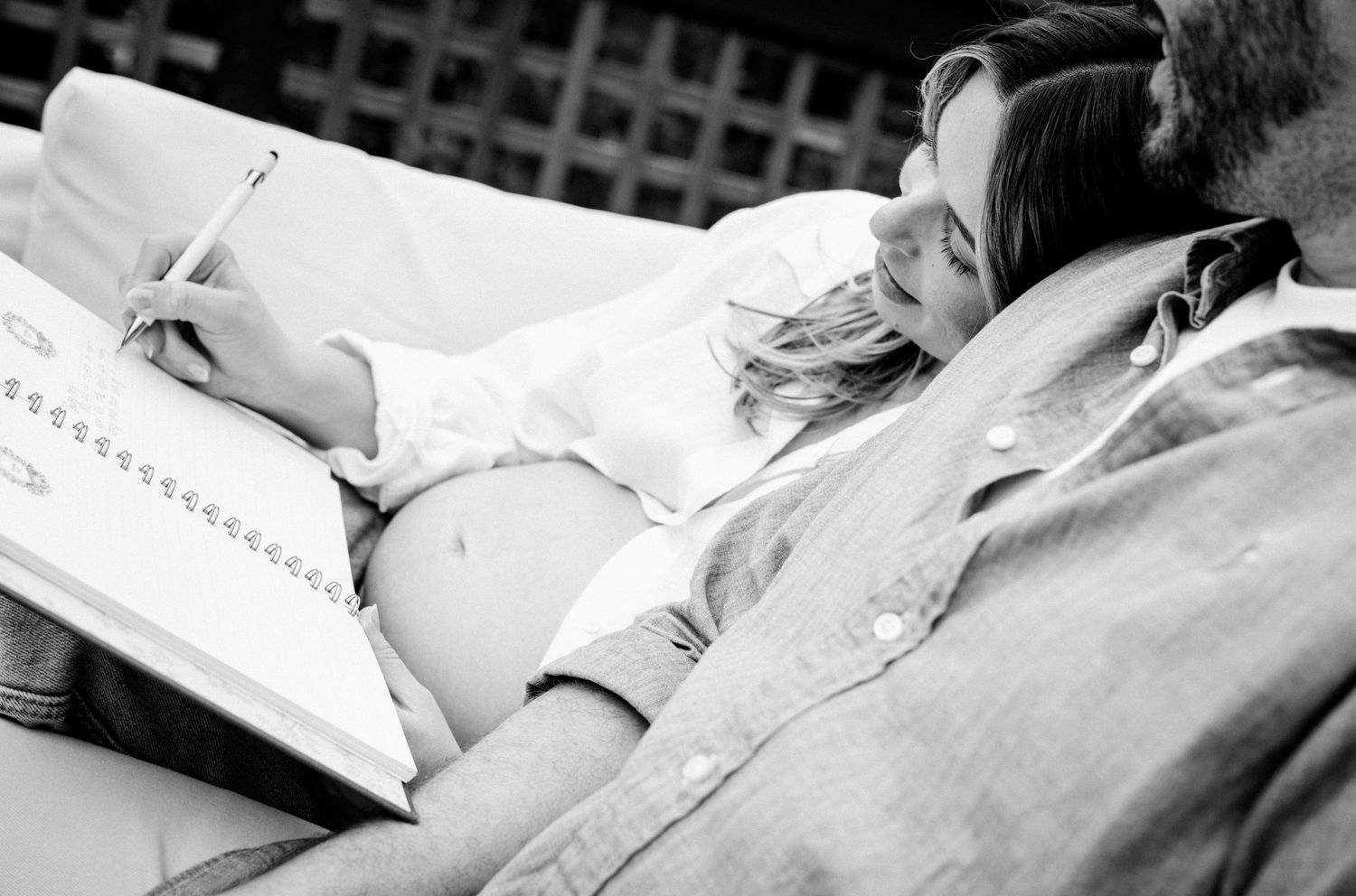 Black and white photo of a pregnant woman writing in a notebook while resting with her partner outdoors.