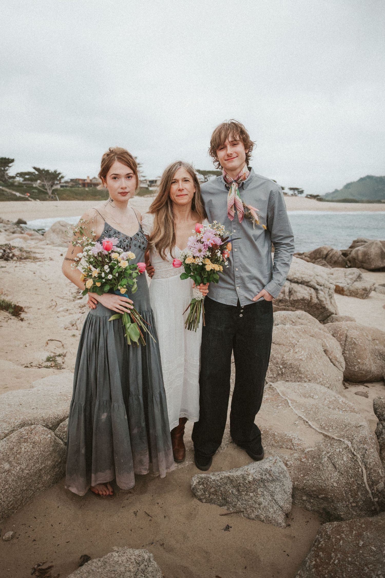 Bride posing with two guests holding colorful bouquets on a rocky beach wedding venue.