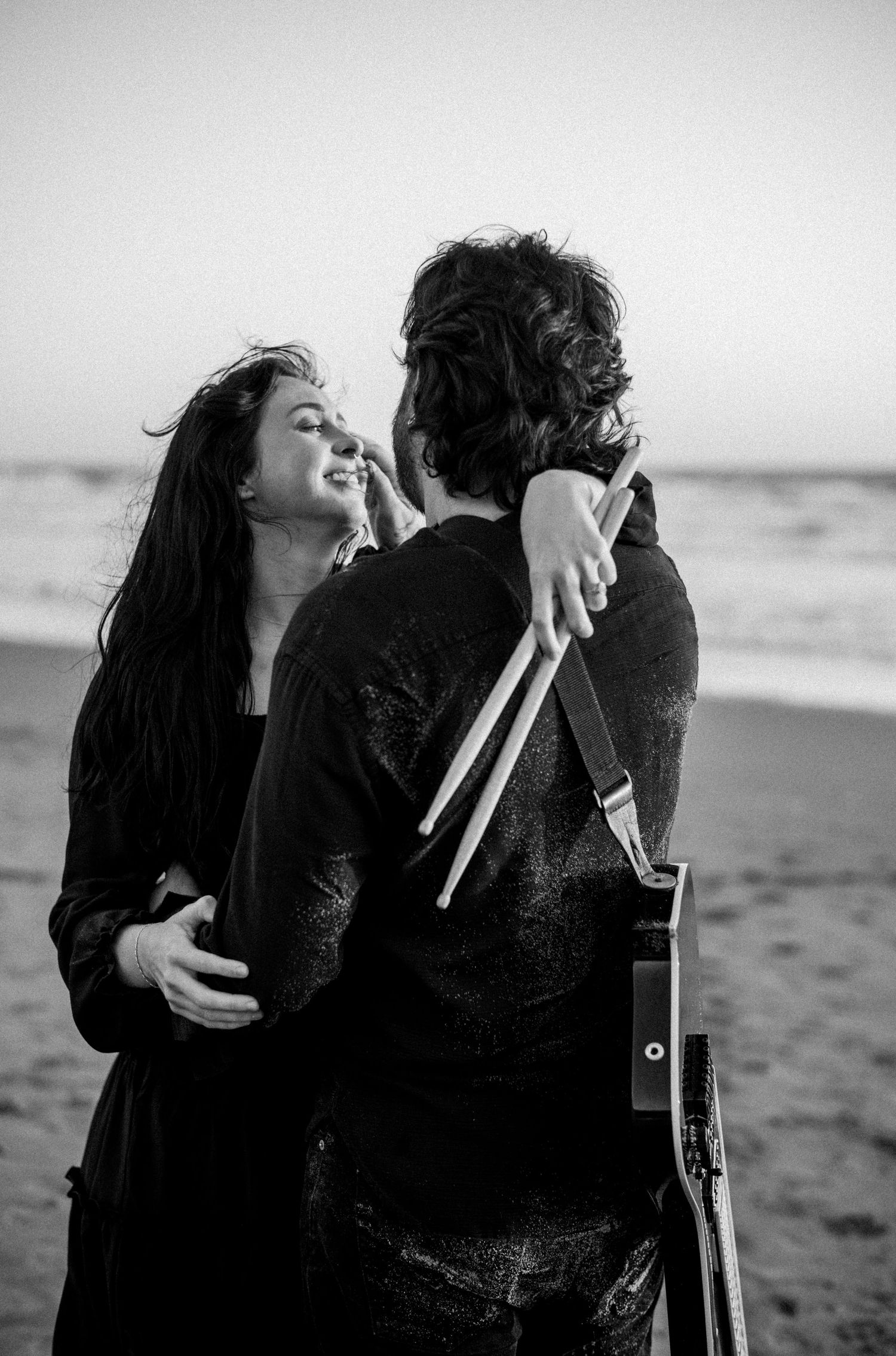 Couple embracing on beach with man holding drumsticks and guitar in black and white.