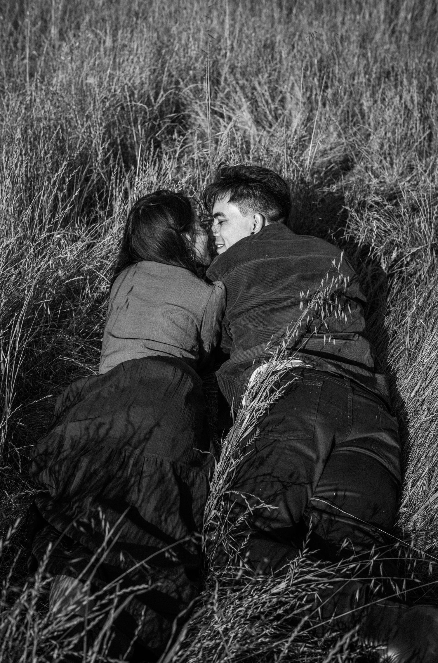 Black and white photo of a couple lying together in tall grass, sharing an intimate moment outdoors.Photography by The Y.K. Studio.