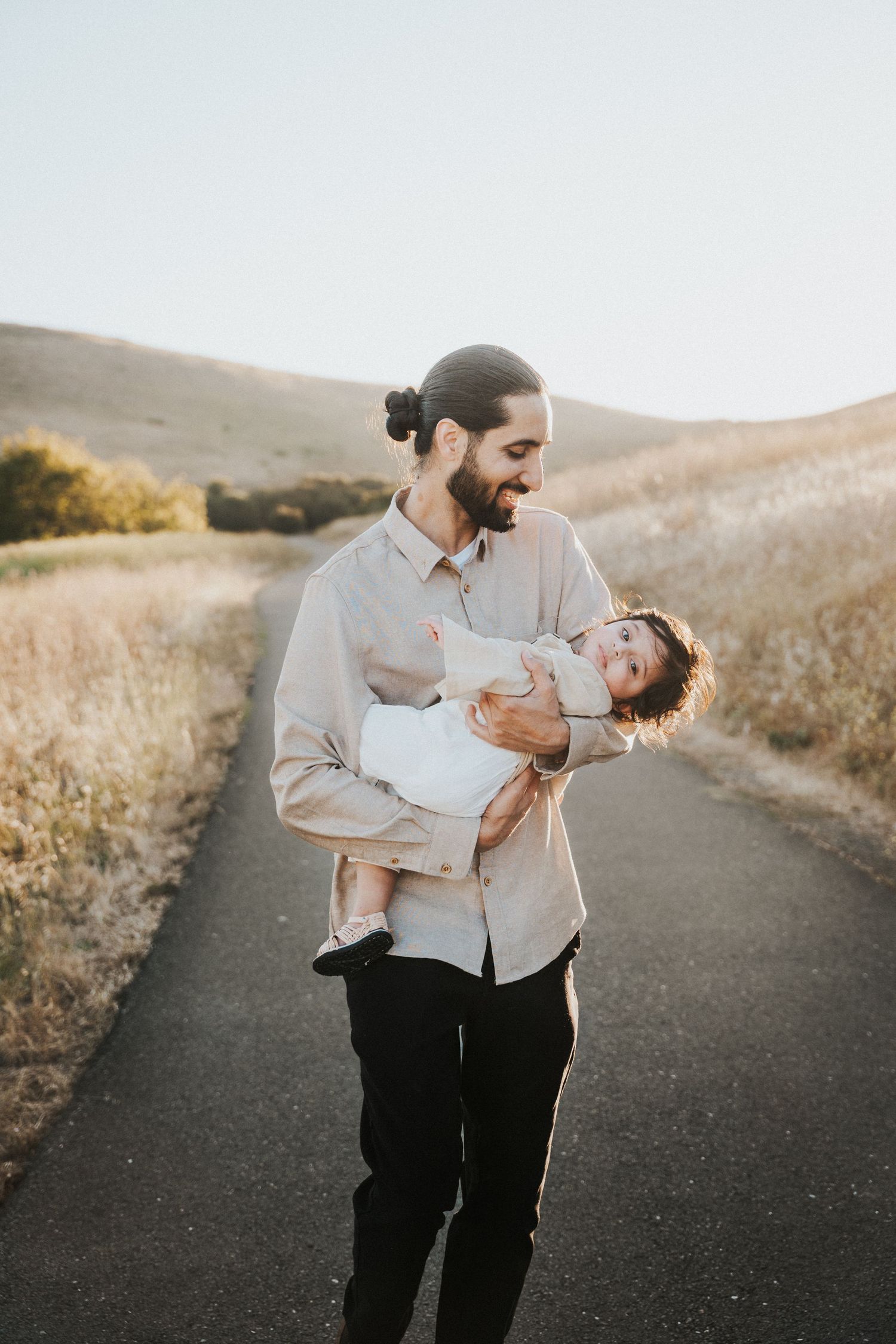 A bearded man with a bun holds a smiling baby on a sunny path surrounded by golden hills.