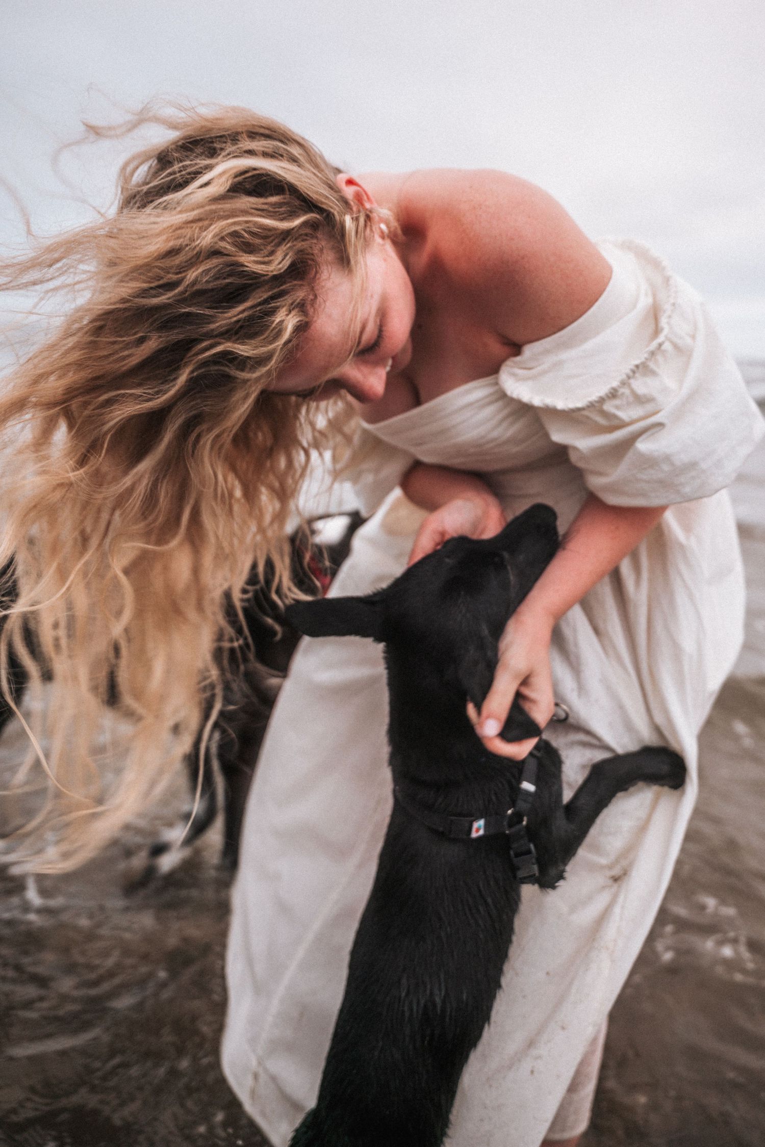 Woman in white off-shoulder dress hugging a wet black dog on a beach.