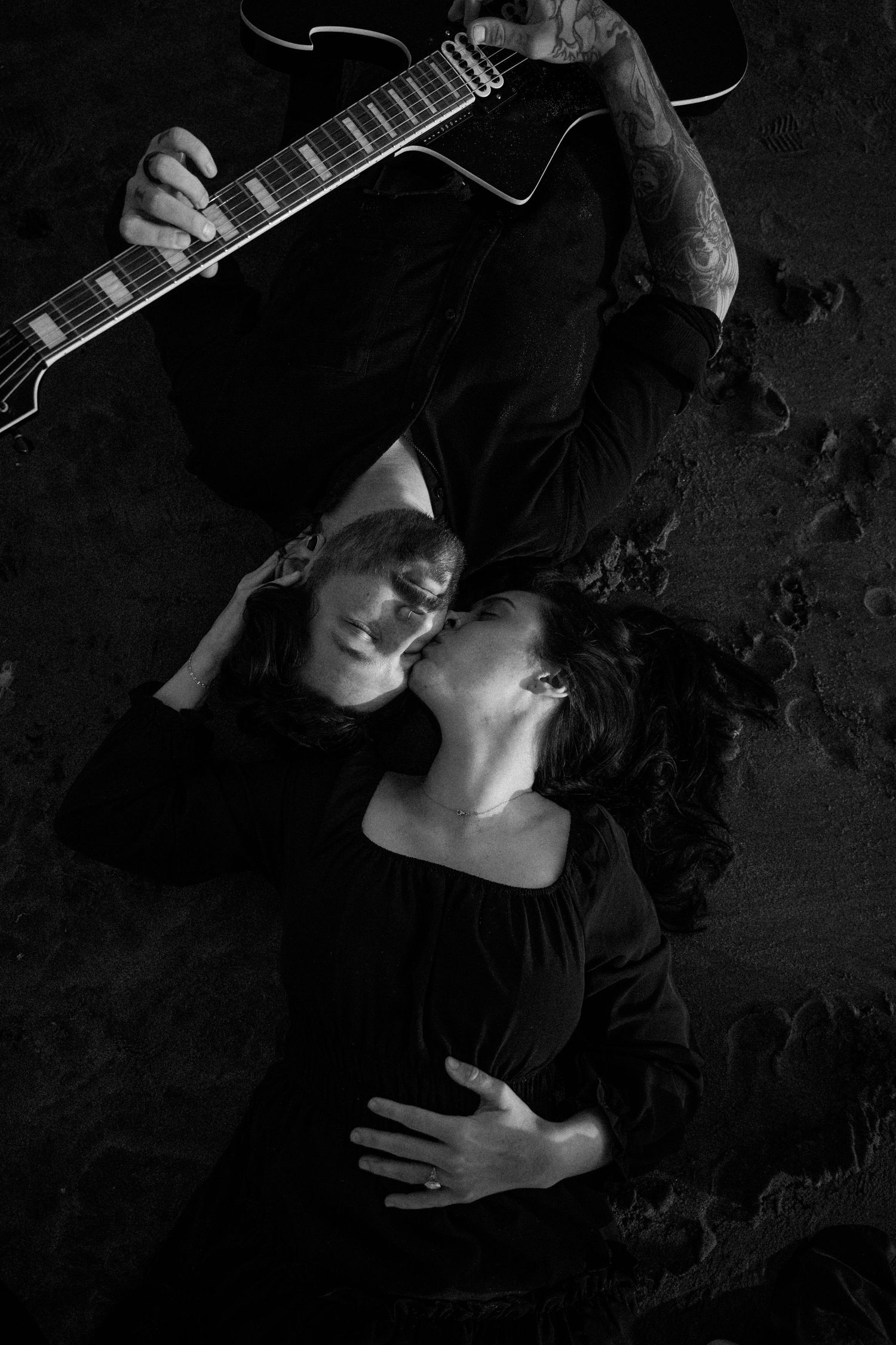Black and white photo of a couple lying on the ground, man holding electric guitar with tattoos visible.