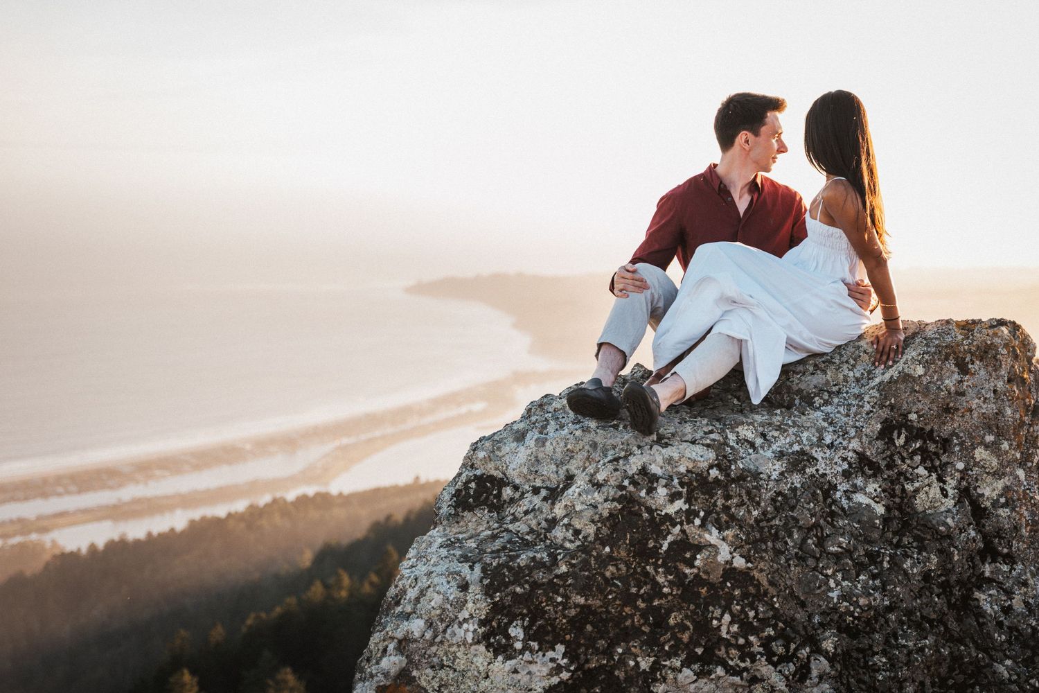 Romantic couple sitting on rocky cliff overlooking coastal ocean view during golden hour sunset.