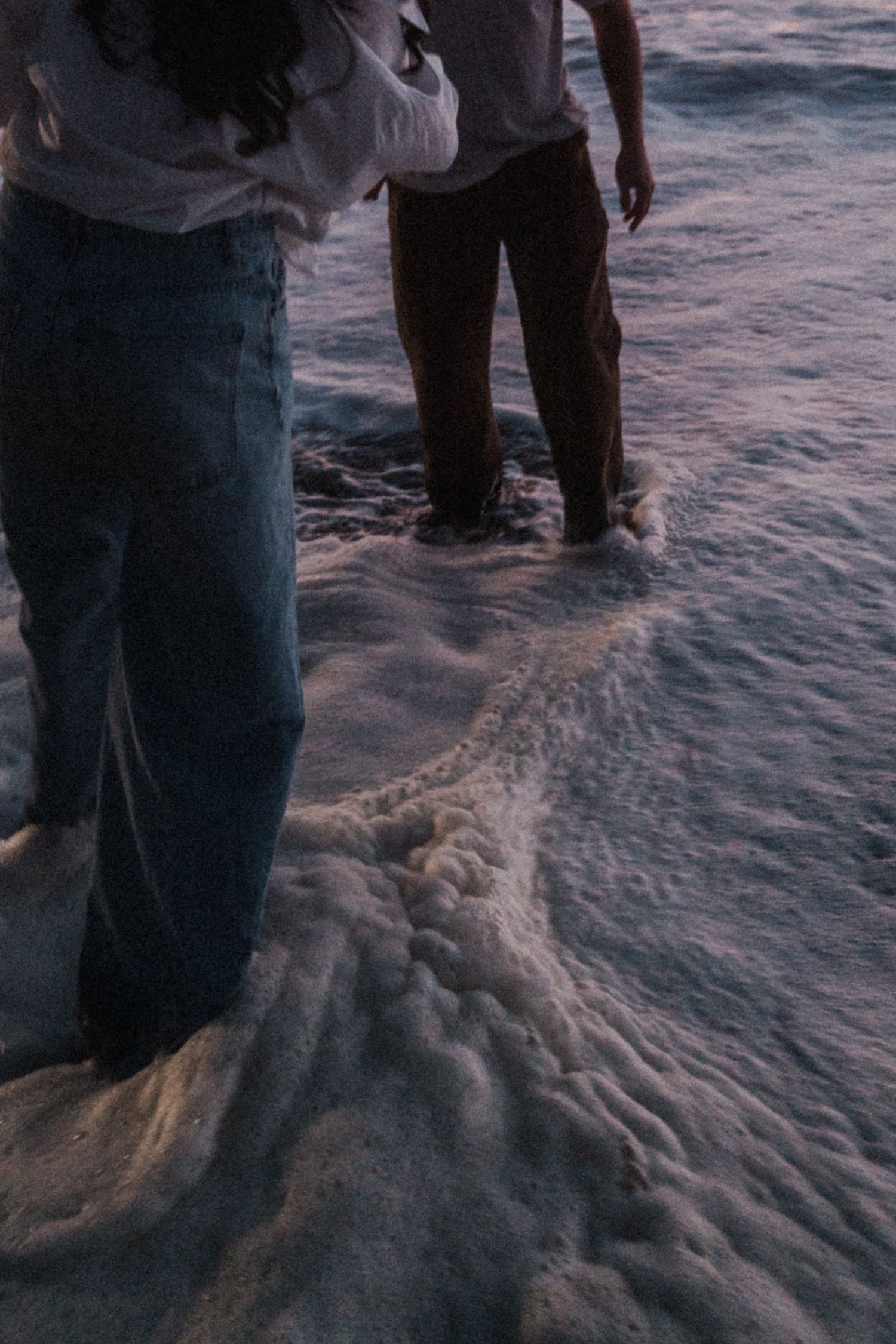 Two people standing at ocean shoreline with waves washing over their feet at dusk, wearing jeans and casual clothes.