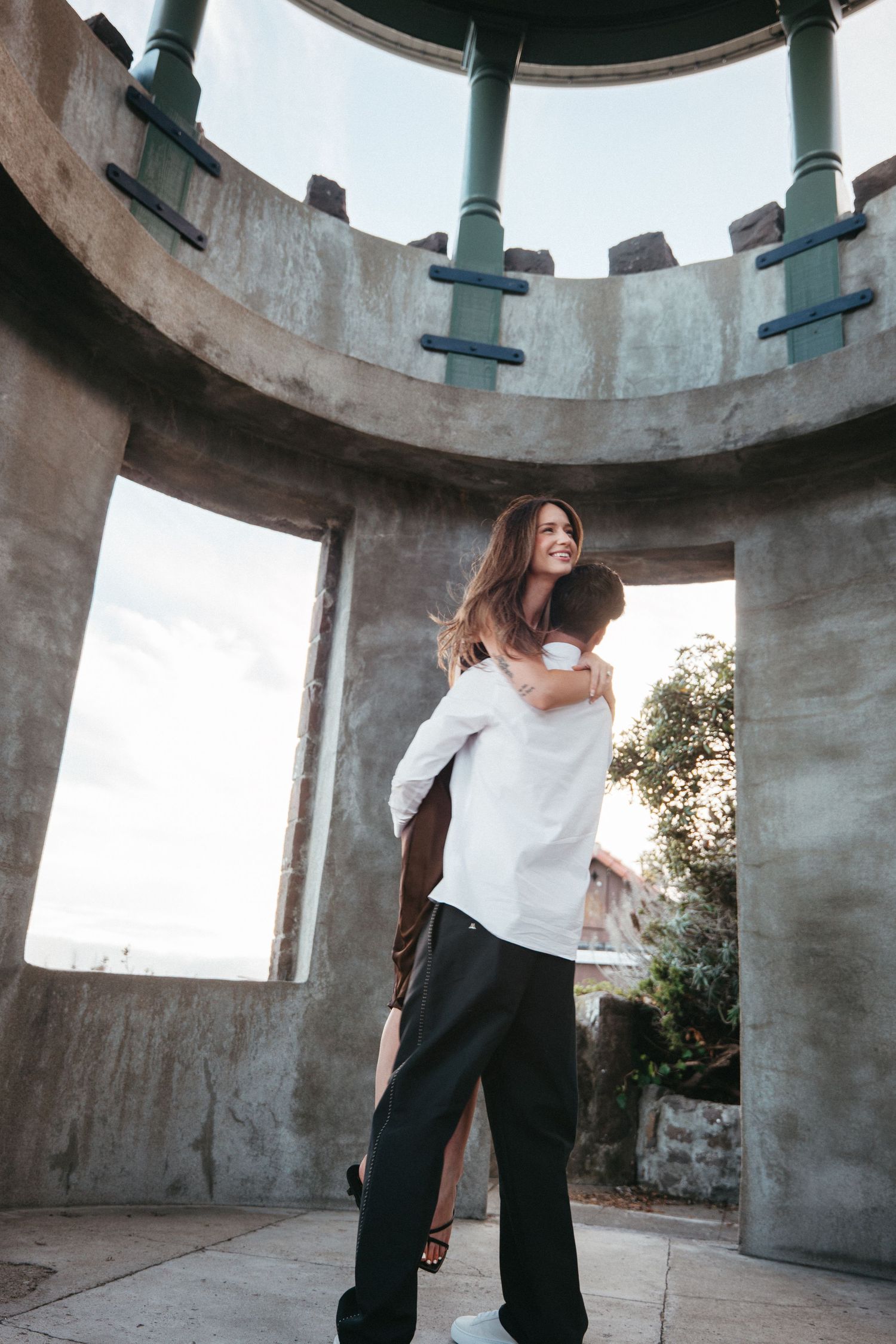 A couple shares a joyful piggyback ride inside a circular stone structure with open archways and green columns.