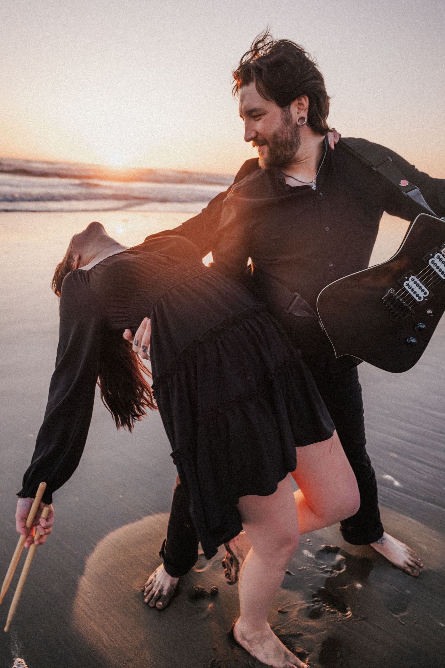 Couple in black outfits dancing on a beach at sunset, man holding woman with guitar on his back.
