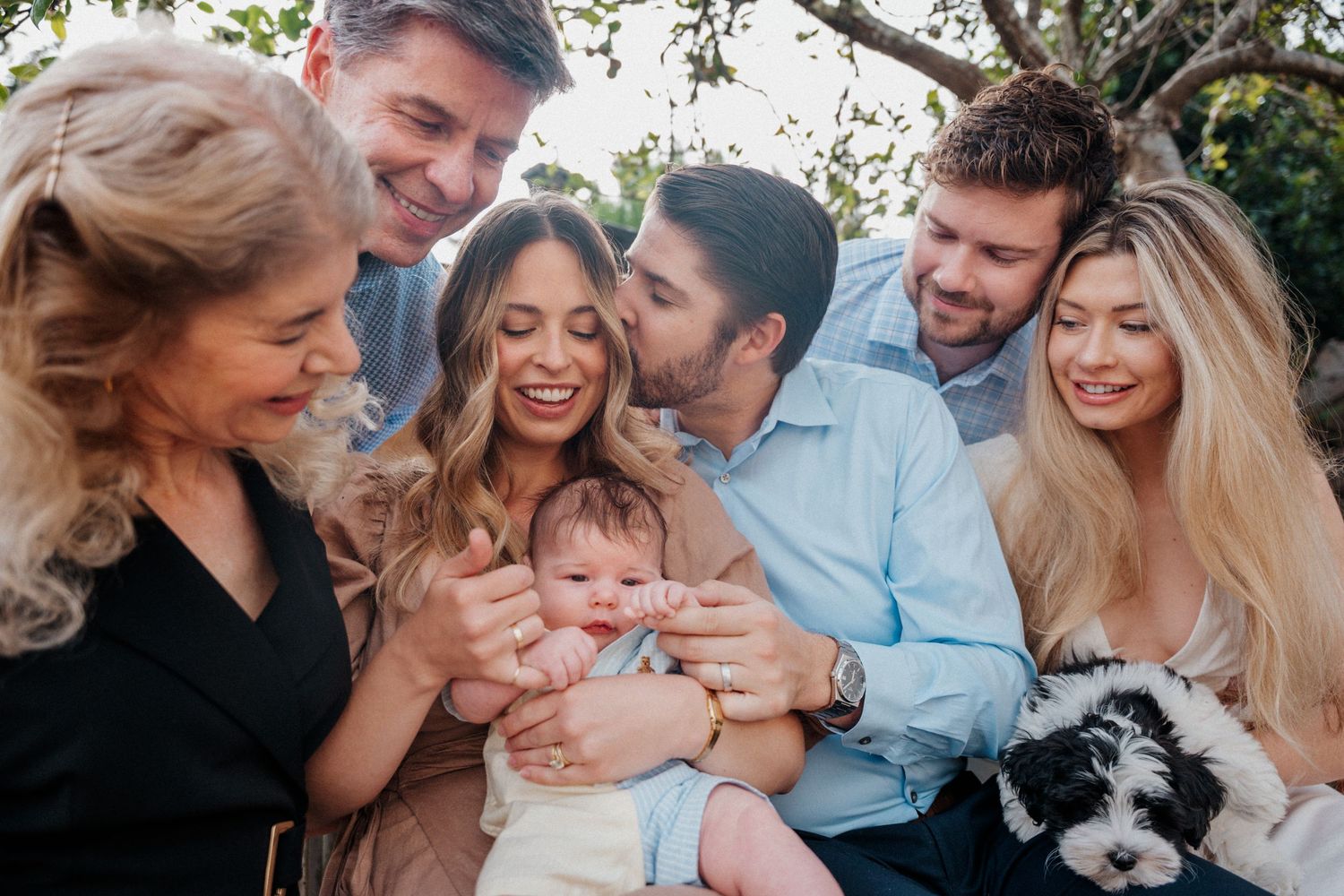 Happy family group photo with parents, grandparents, and siblings gathered around a newborn baby and puppy outdoors.