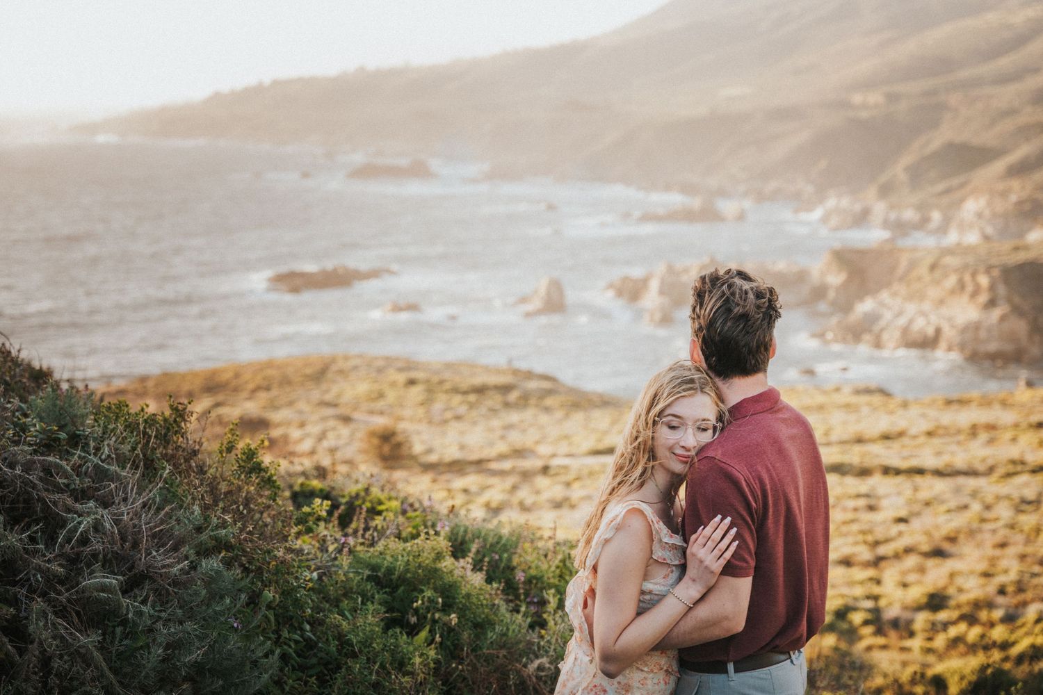Couple embracing on a scenic coastal cliff overlooking rocky shores and golden hills at sunset.