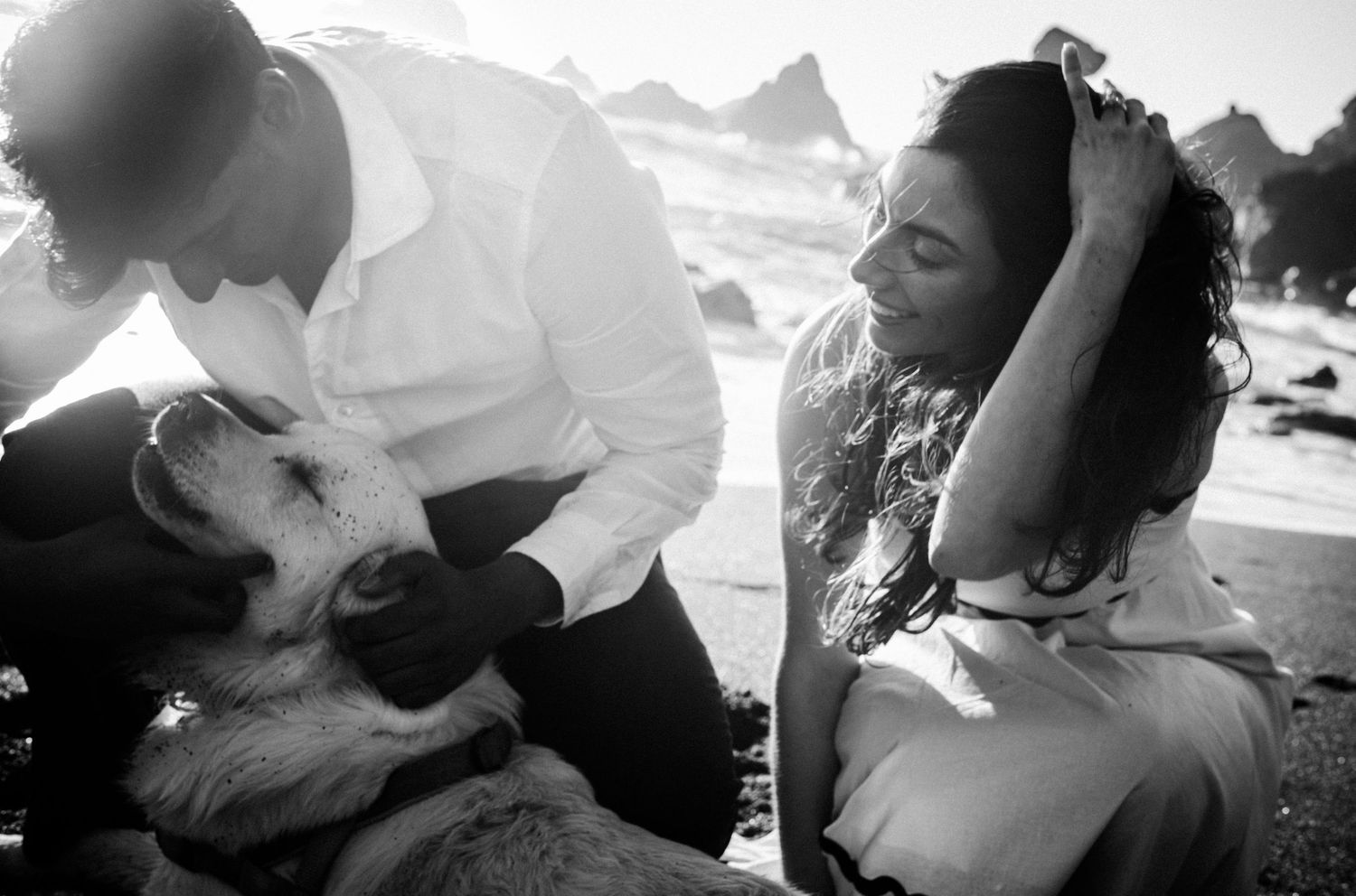 Black and white photo of a couple petting a dog on a beach.