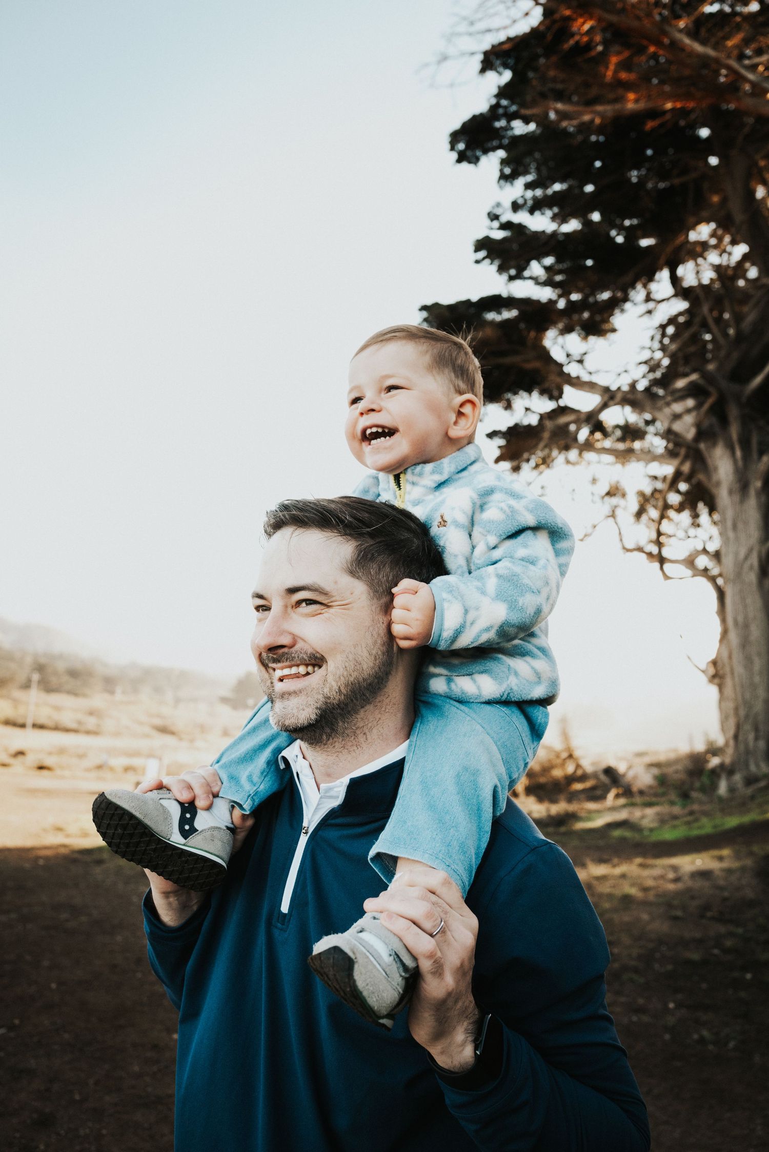 A smiling father carries a laughing toddler on his shoulders outdoors near a tree on a sunny day. Mori Point, Pacifica.