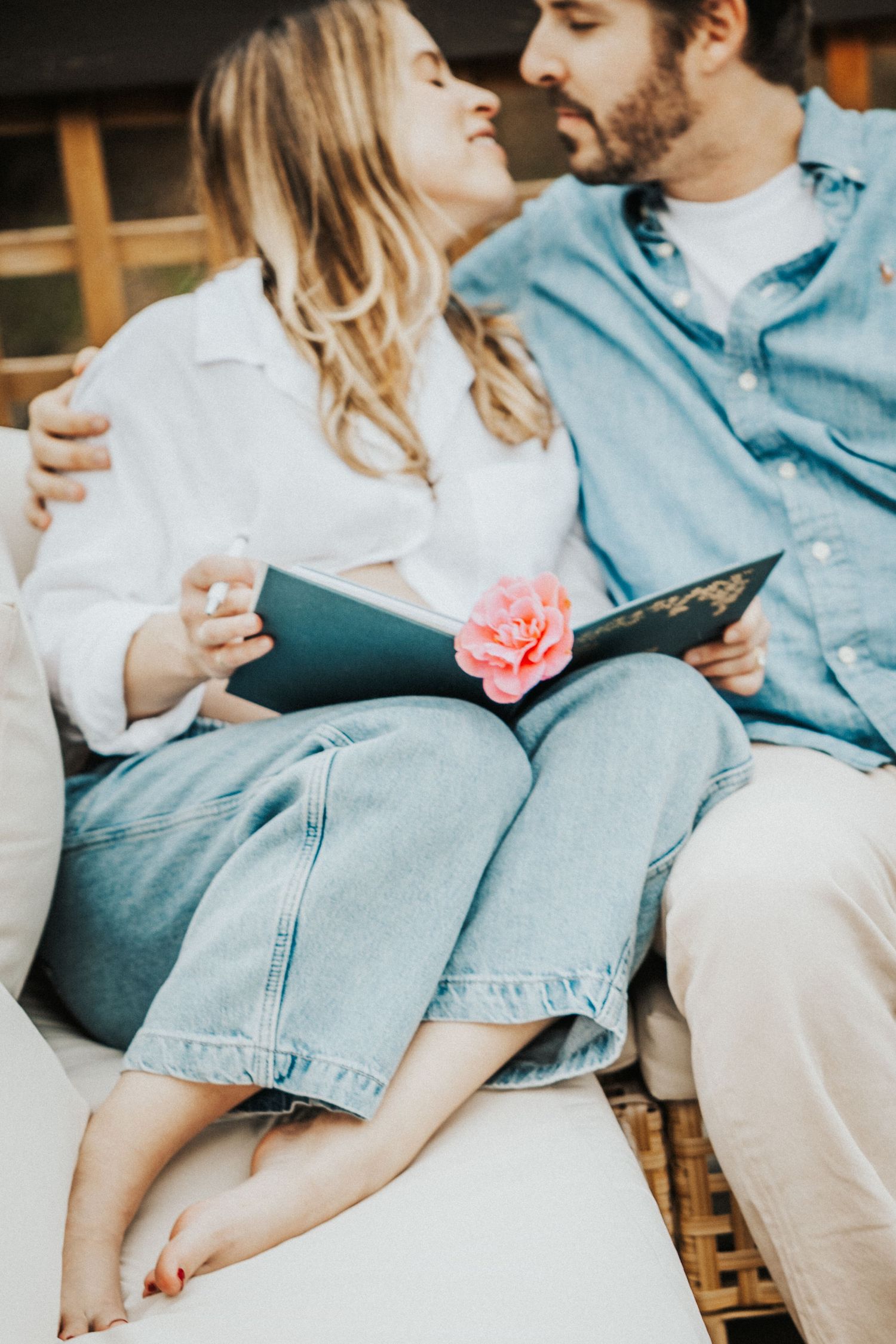 Couple sitting together on a couch, sharing a book with a pink rose, smiling at each other in casual denim outfits.