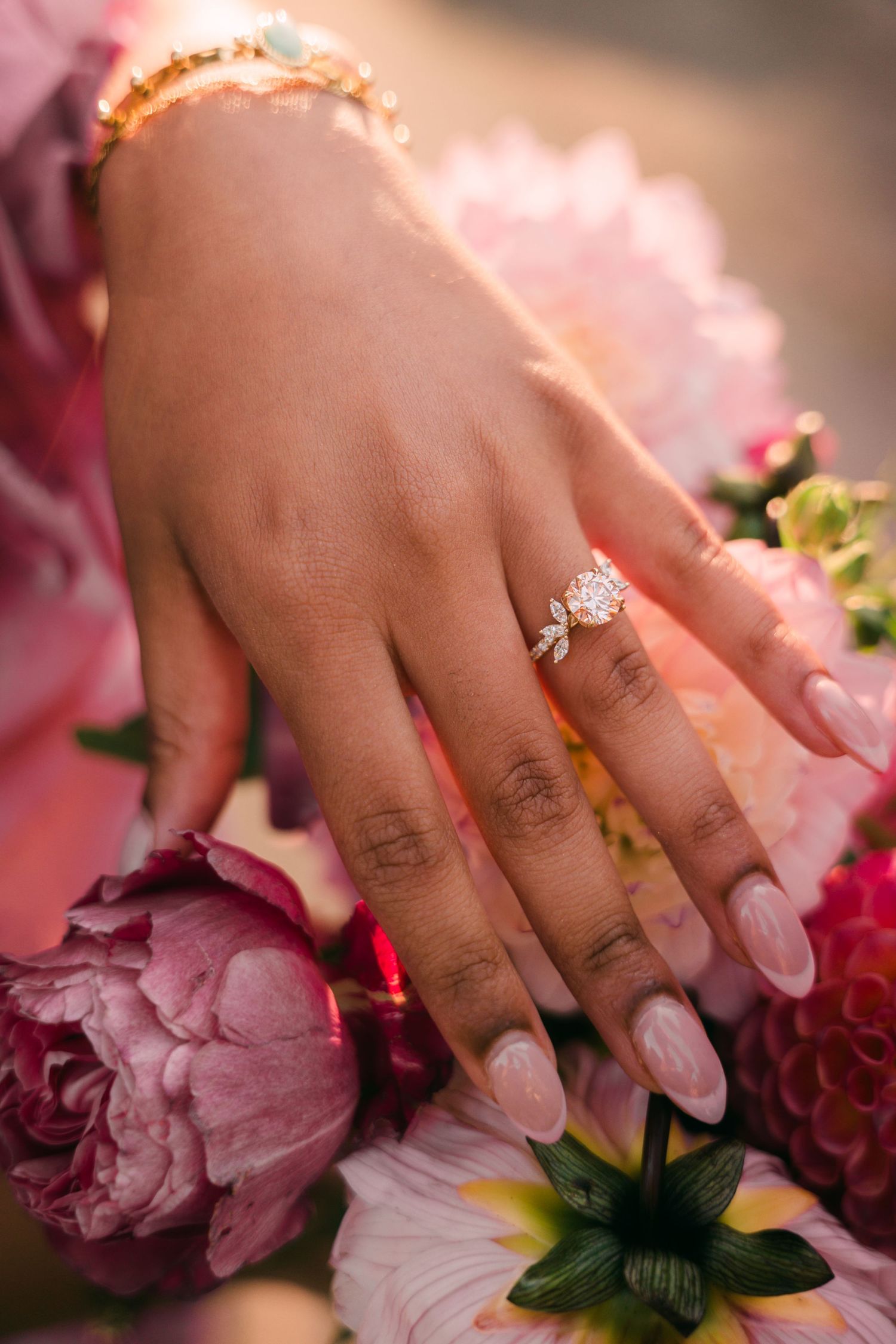 Close-up of a bride's hand wearing a diamond engagement ring with leaf details, resting on pink peonies.