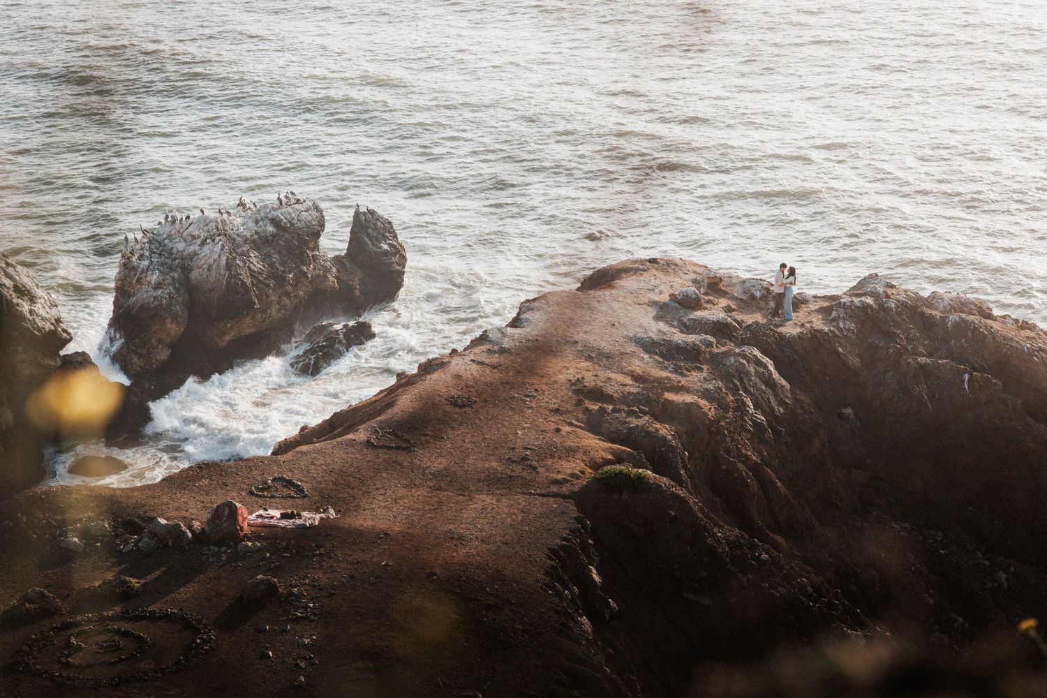 Couple standing on rocky coastal cliffs overlooking crashing ocean waves at golden hour.