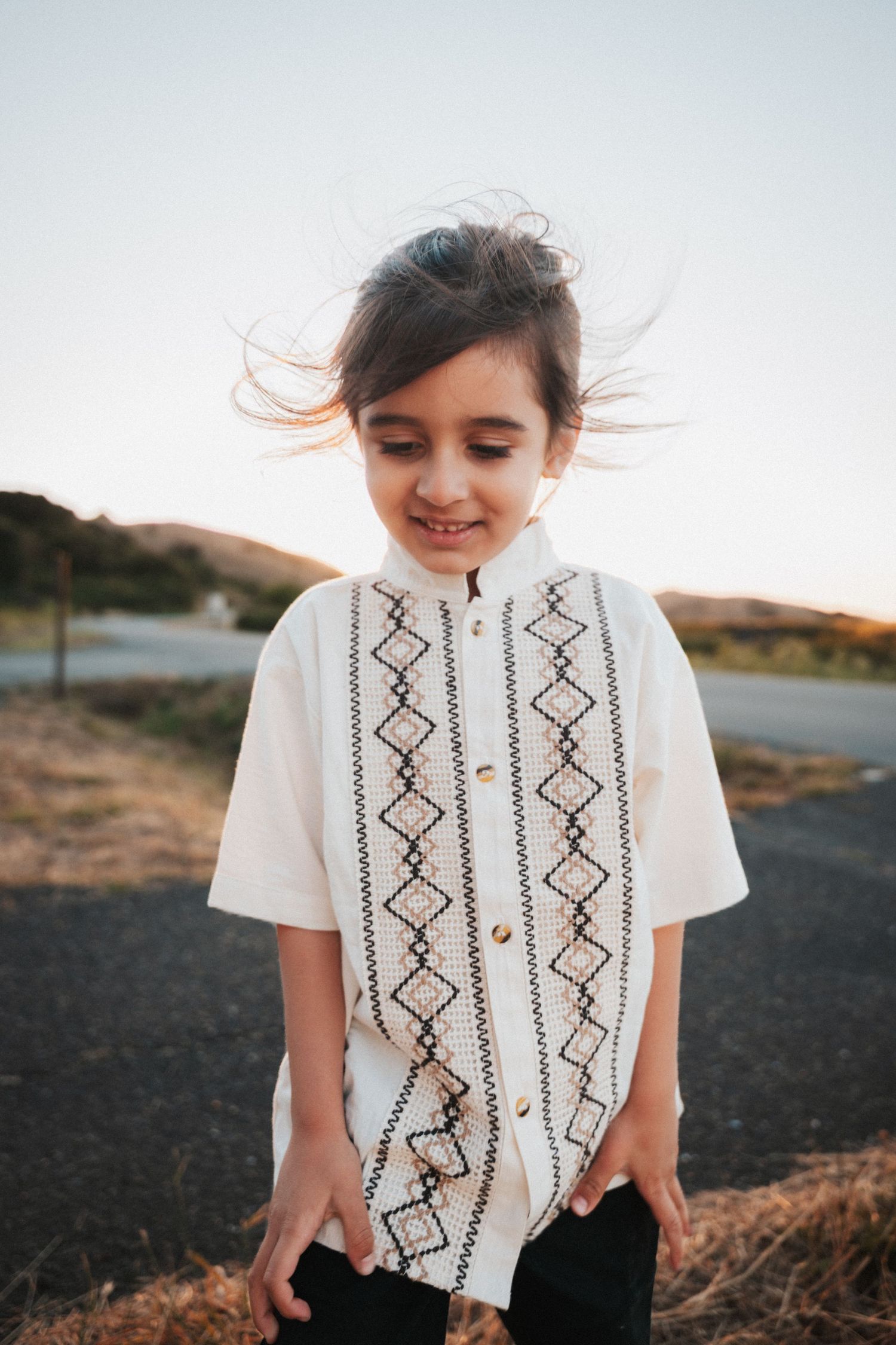 A young girl in a white embroidered vest smiles outdoors at golden hour, her hair blowing in the wind.