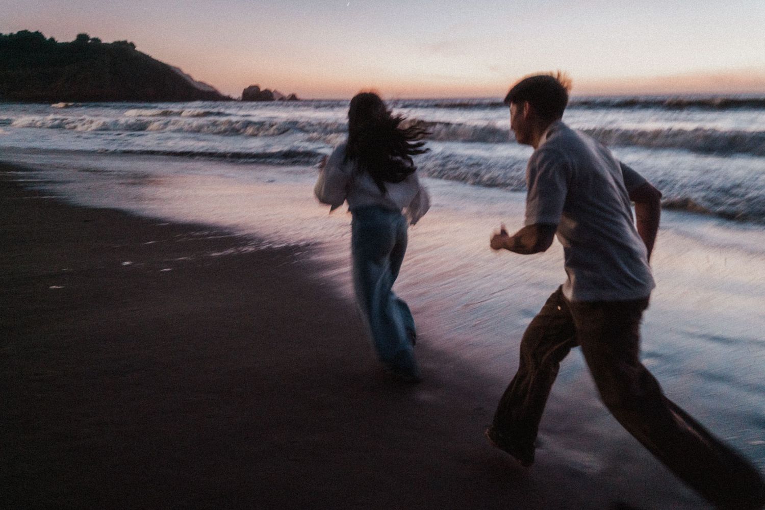 Two people running joyfully along a sandy beach at dusk, with ocean waves and a colorful sunset sky in the background.