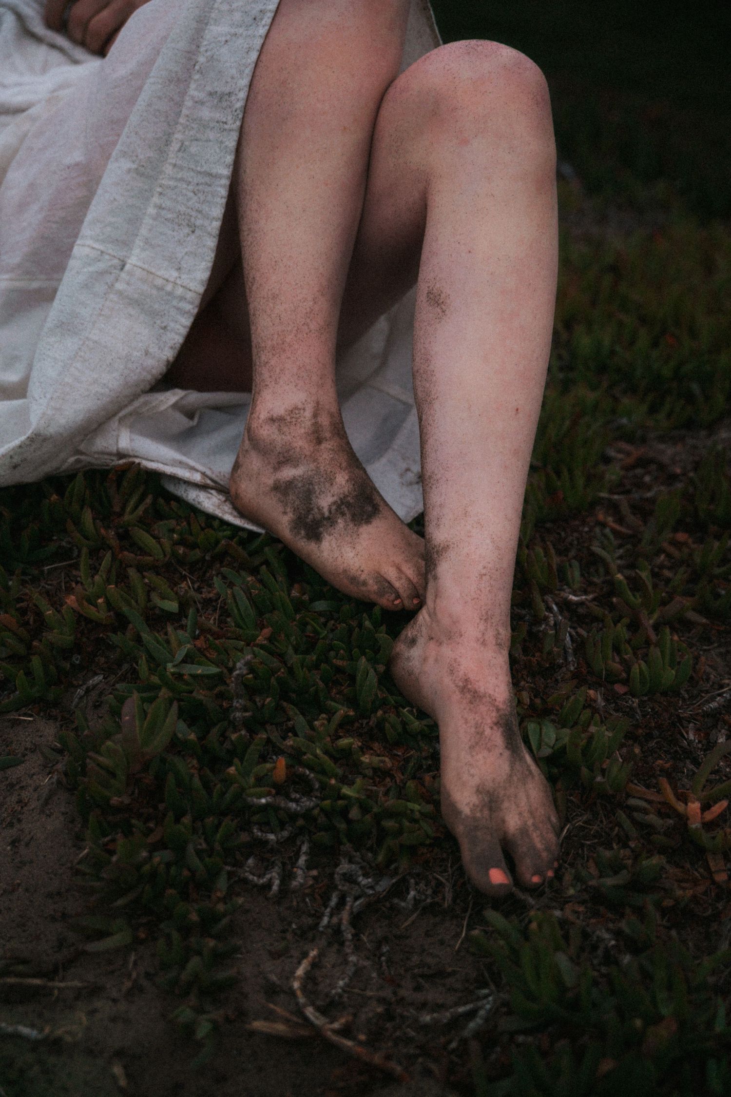 Close-up of a woman's bare feet and legs covered in dark sand while sitting on grass outdoors.
