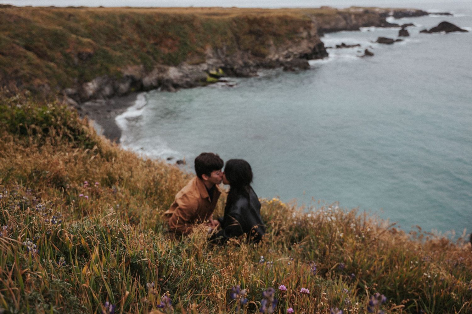 Couple sharing a romantic kiss on a coastal cliffside meadow with ocean waves in the background.