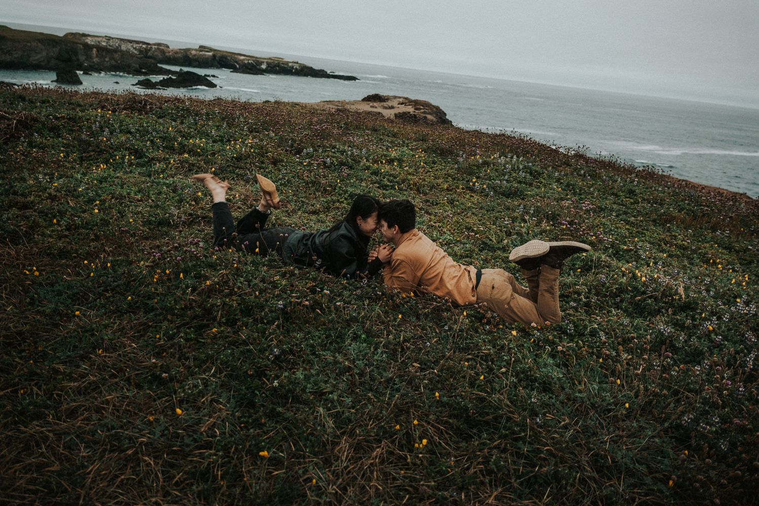 Couple lying on a grassy coastal hillside sharing a tender moment with the ocean in the background.