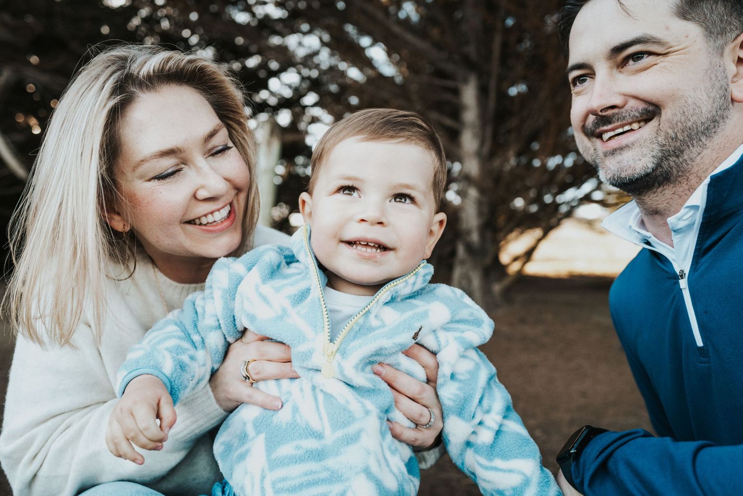 Happy family with smiling parents holding their cheerful baby outdoors near trees in a warm, candid portrait.