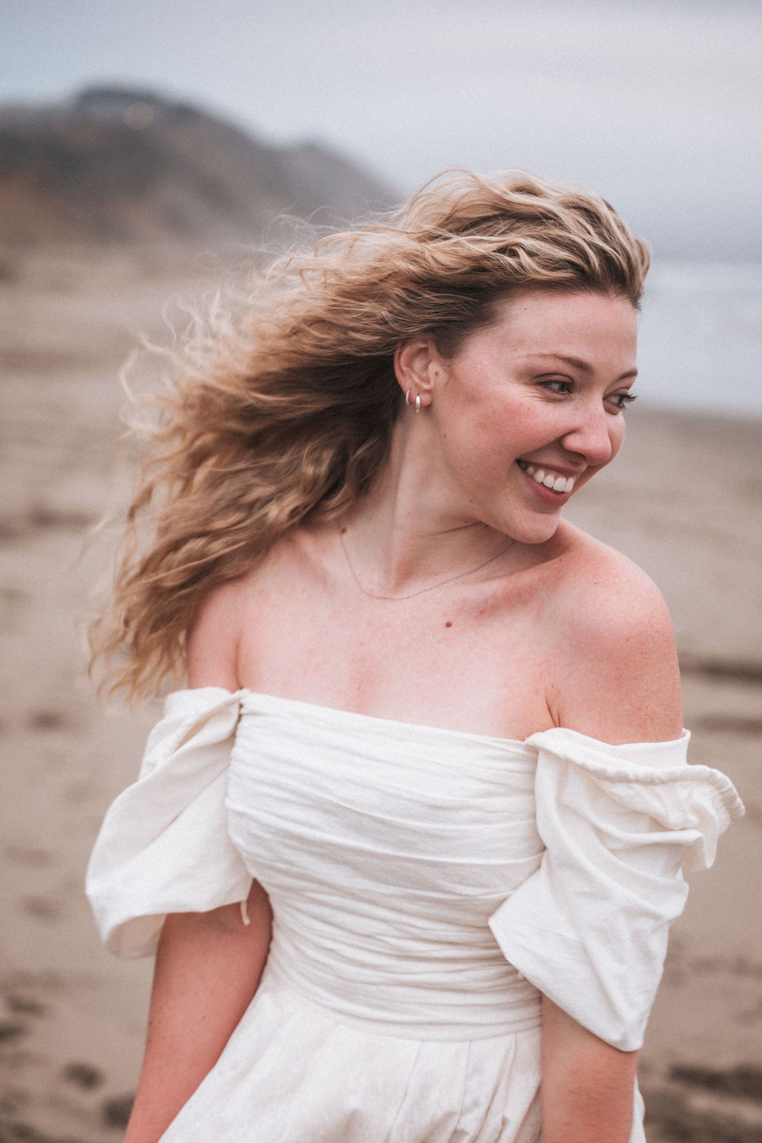 Smiling blonde woman in white off-shoulder dress with wind-blown hair on a sandy beach.