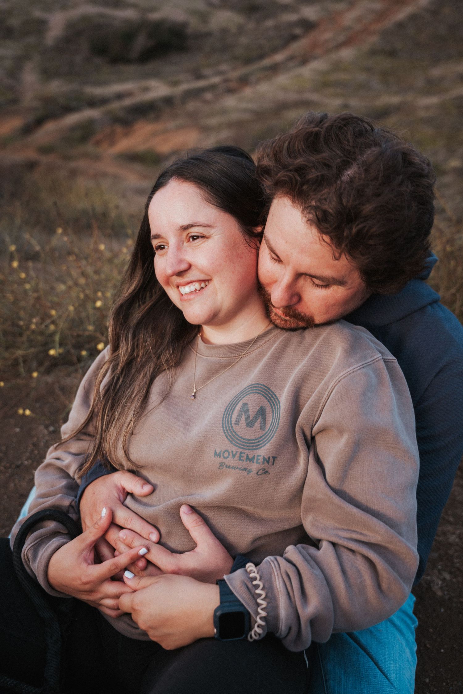 A couple shares a tender embrace outdoors, man kissing woman's cheek as she smiles, wearing a Movement sweatshirt.