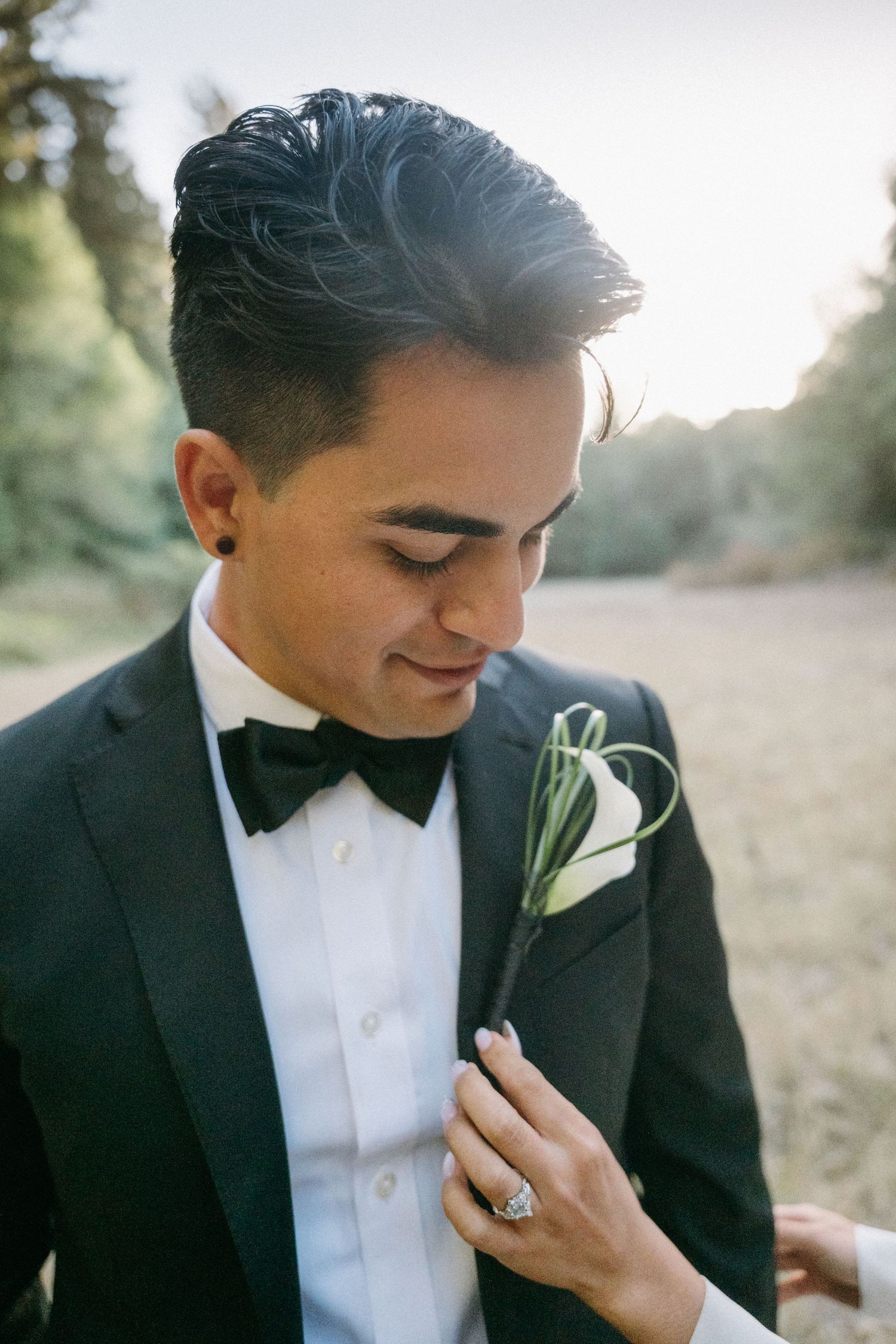 Close-up of groom in black tuxedo and bow tie looking down as bride pins a floral boutonniere to his lapel outdoors.