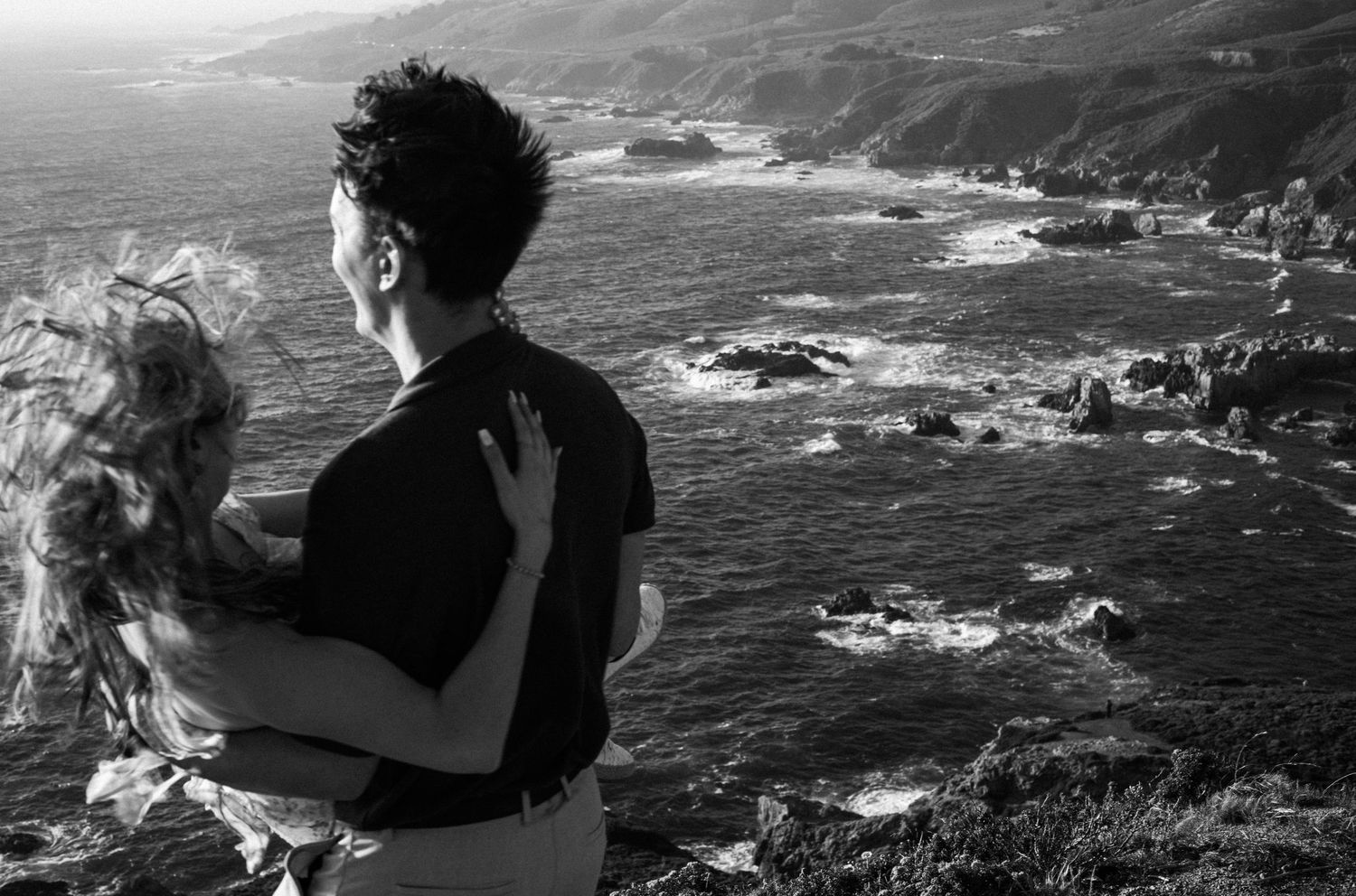 Black and white photo of a couple embracing on a coastal cliff overlooking rocky Pacific Ocean shoreline.