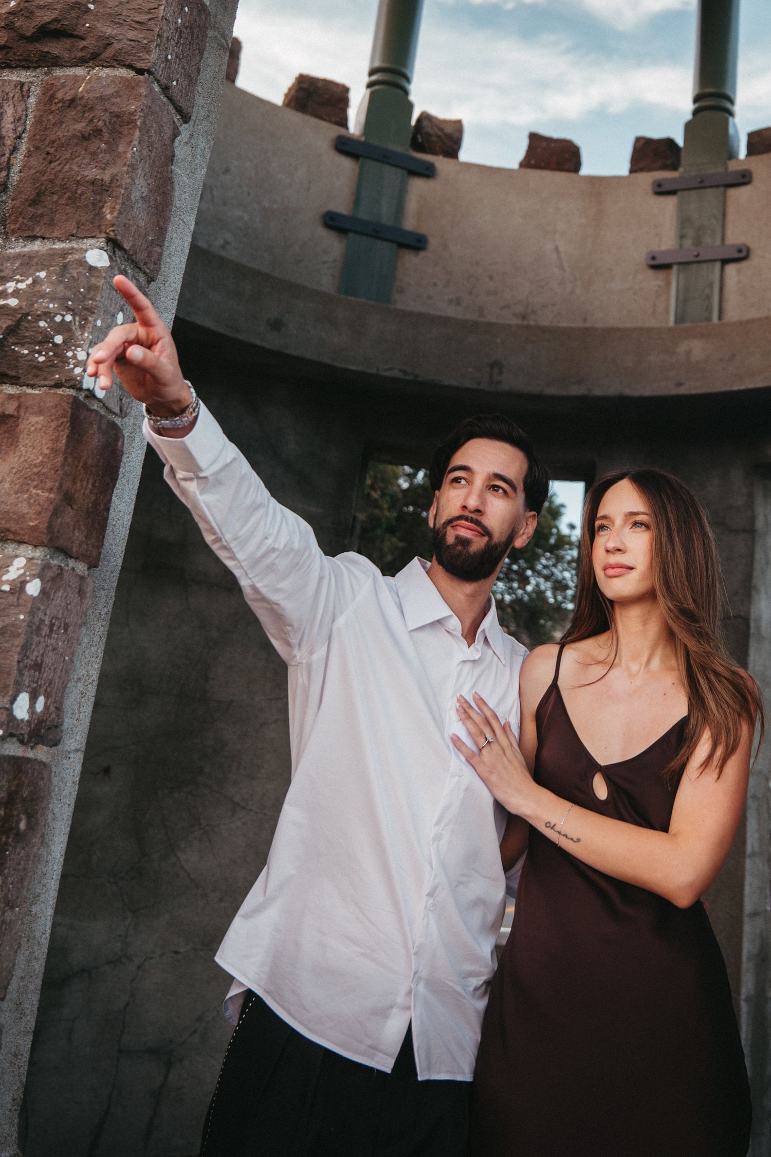A bearded man in a white shirt points into the distance while a woman in a brown dress leans against him outdoors.