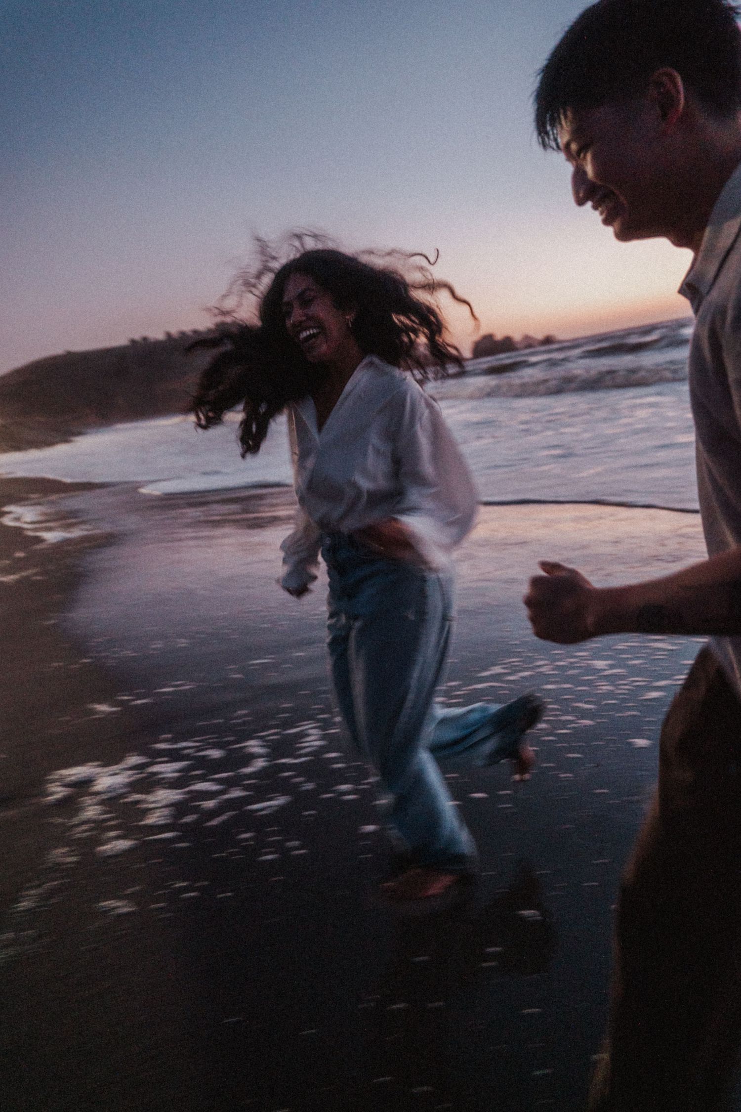 Couple laughing and running on a wet beach at sunset, woman in white top and blue jeans with hair blowing in wind.