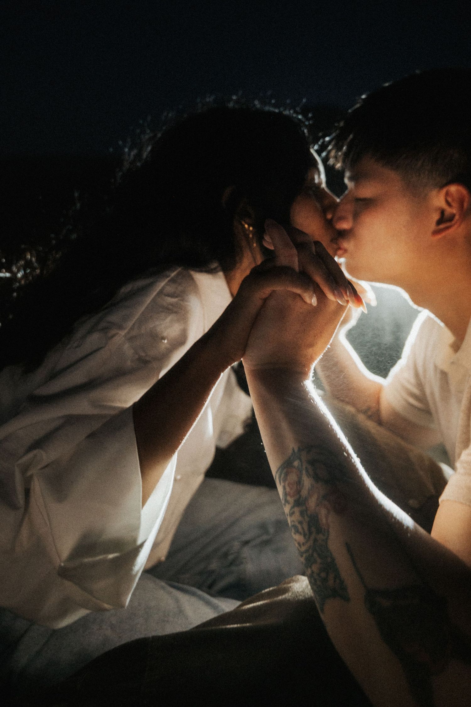 Couple sharing a romantic kiss while holding hands, illuminated by warm light against a dark background.