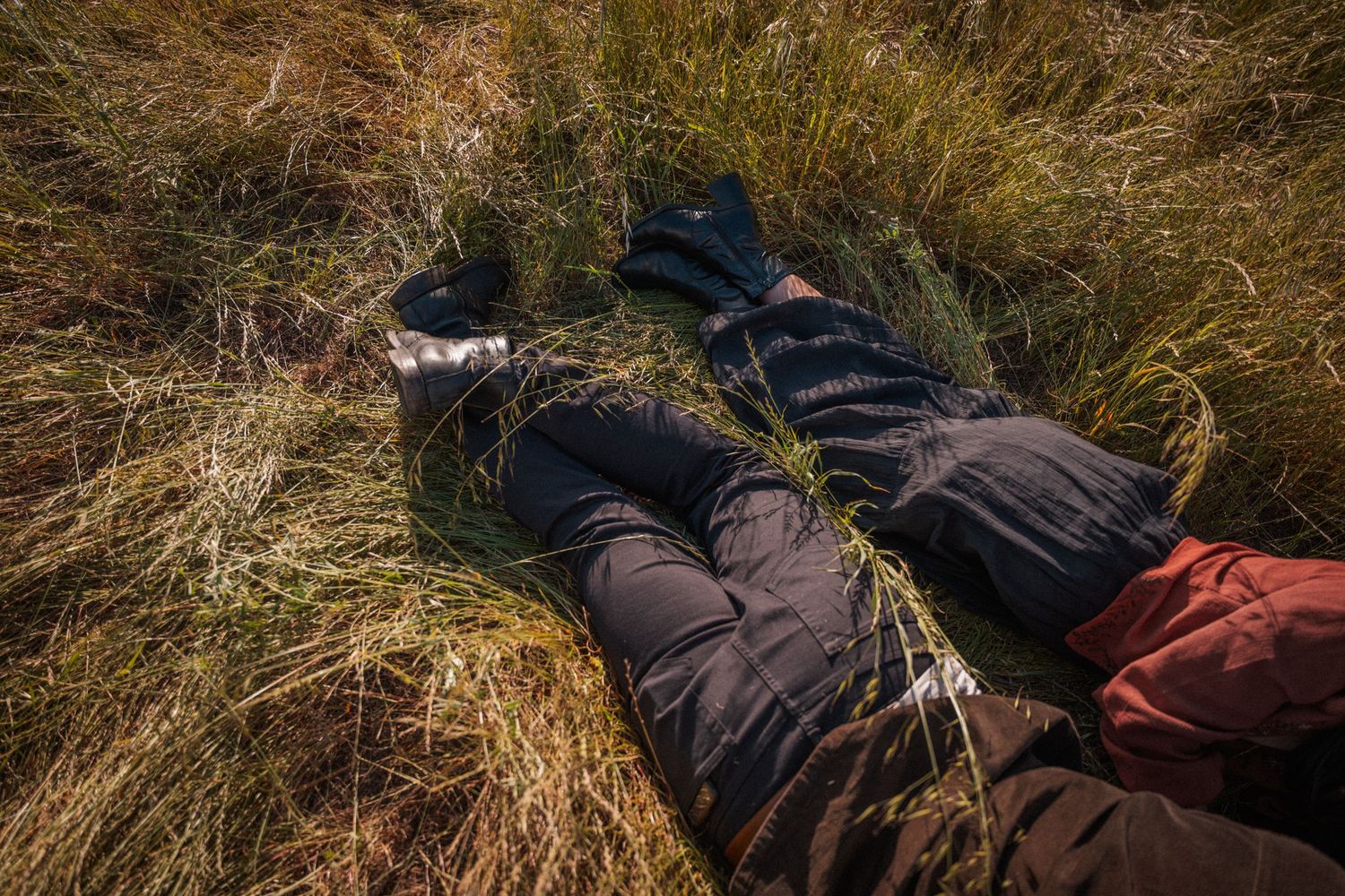 Two people lying in tall golden grass wearing dark outdoor clothing and boots on a sunny day.